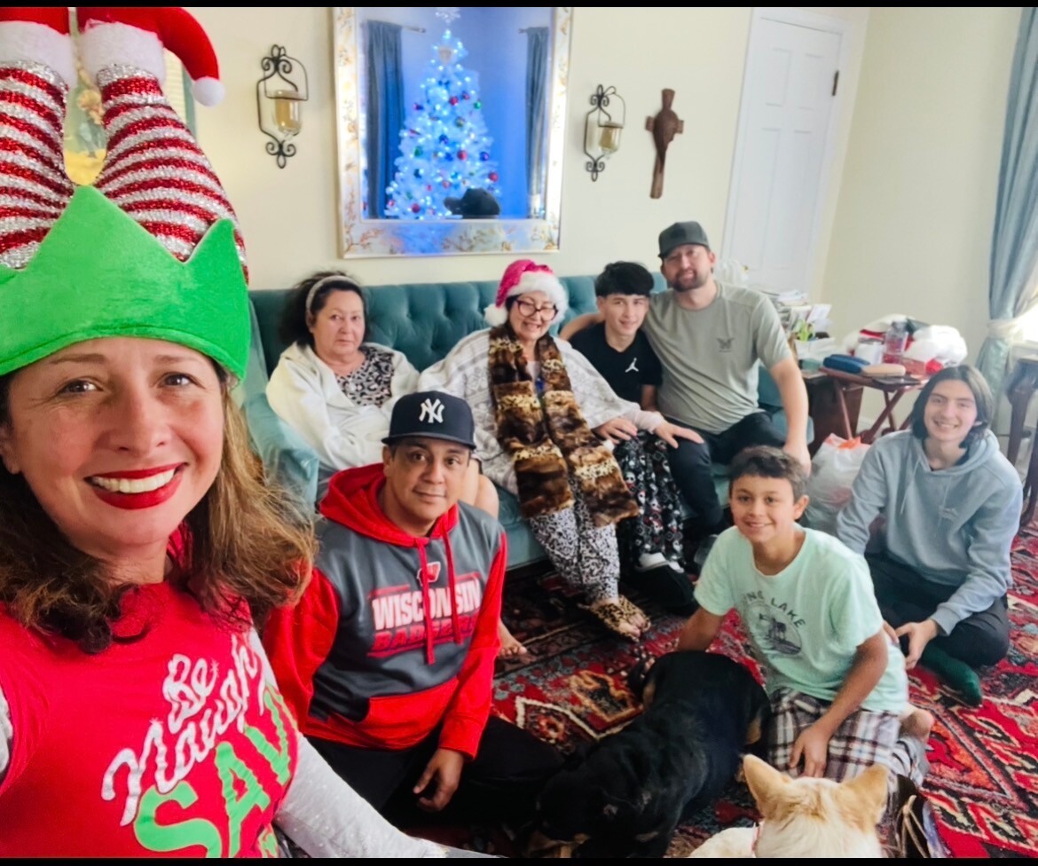 Woman in an elf hat takes a selfie with a multi-generational Christmas gathering on a sofa and carpeted floor.