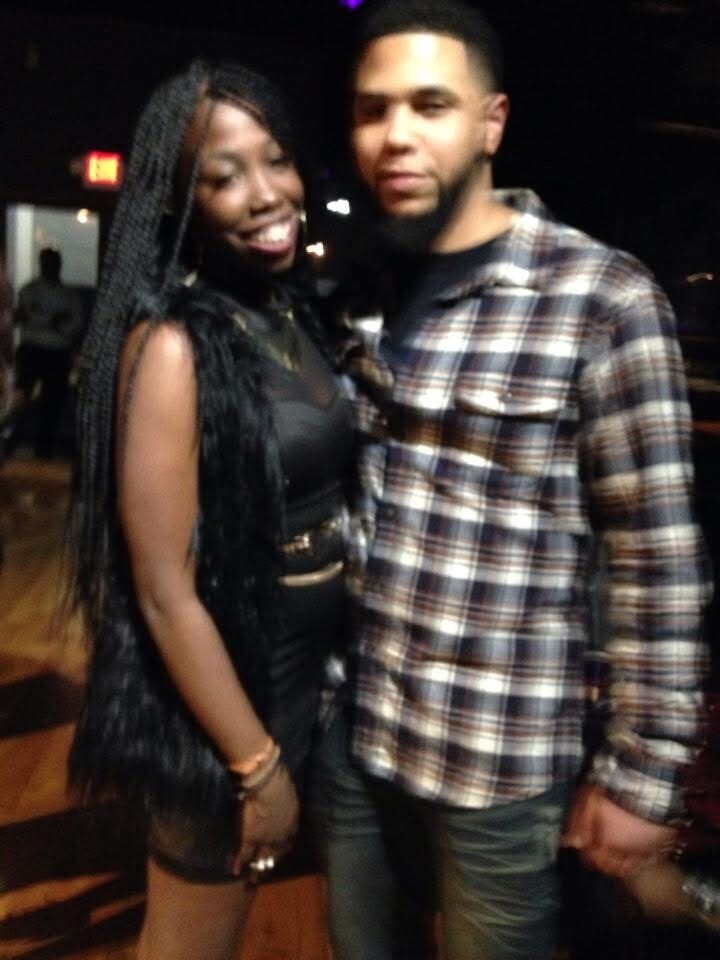 Smiling couple posing together in a dim venue; woman with long braids in black outfit, man in a plaid shirt.