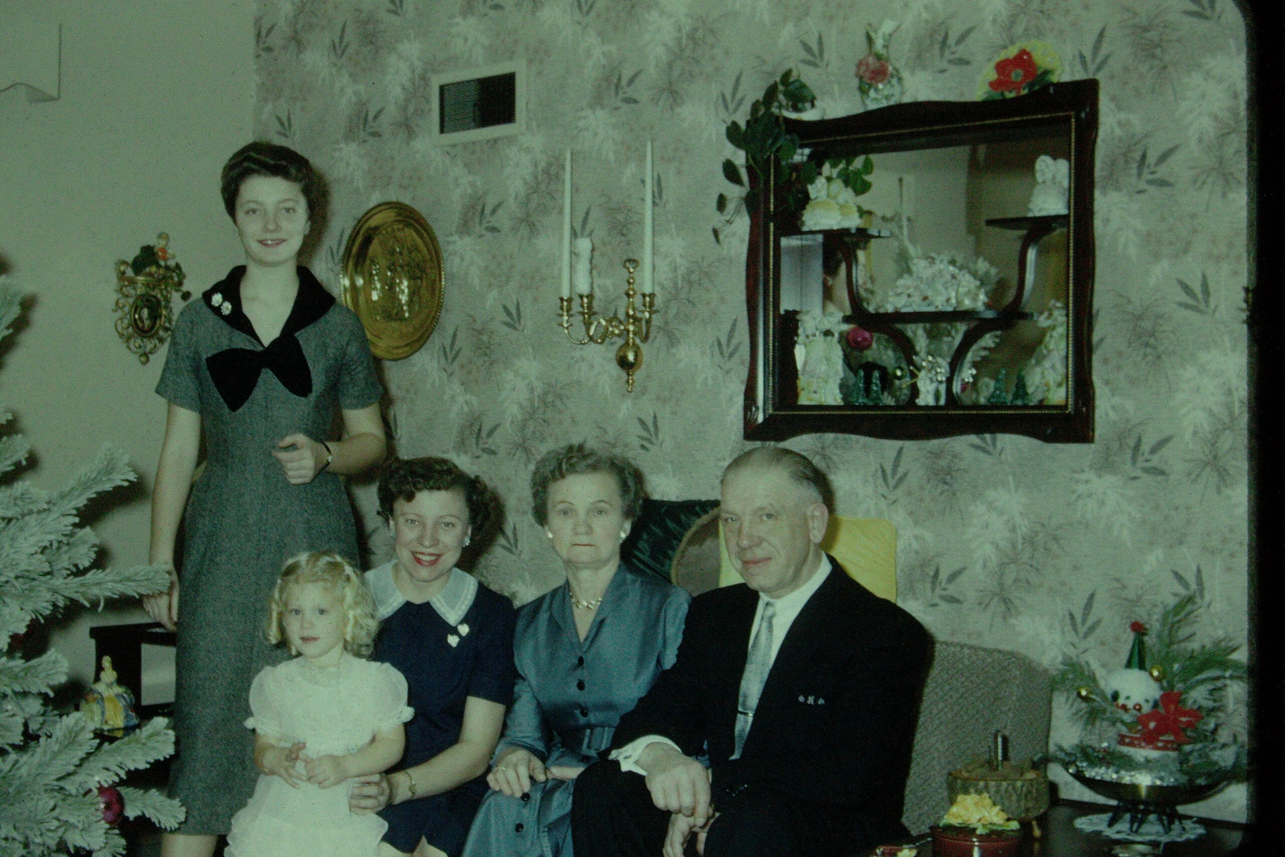 Family Christmas photo: a girl in a white dress holds a cake while relatives pose on a sofa in a living room with a decorated tree nearby.