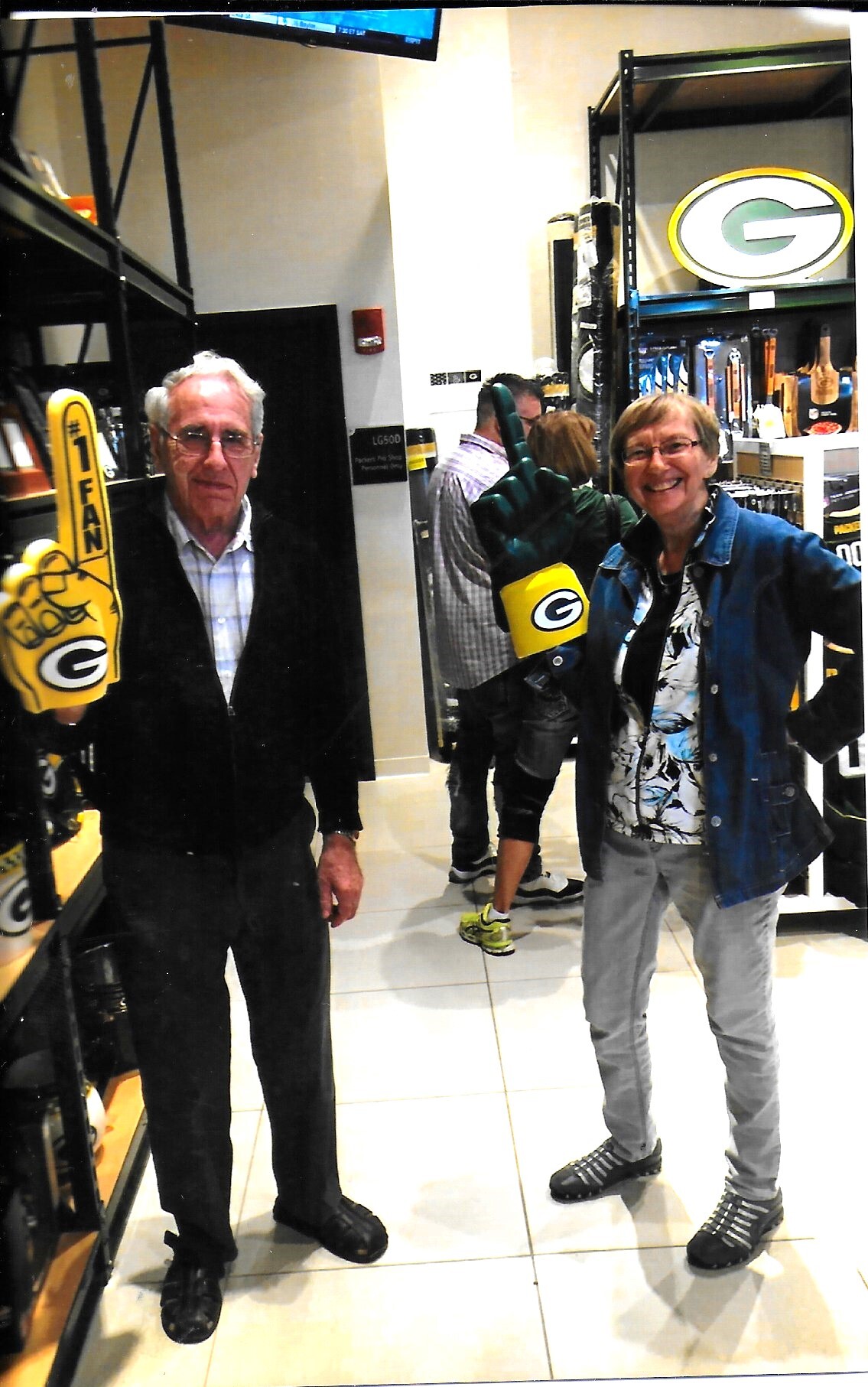 Two smiling adults pose in a sports store, wearing Packers gear and oversized yellow foam fingers with a green G logo on shelves behind them