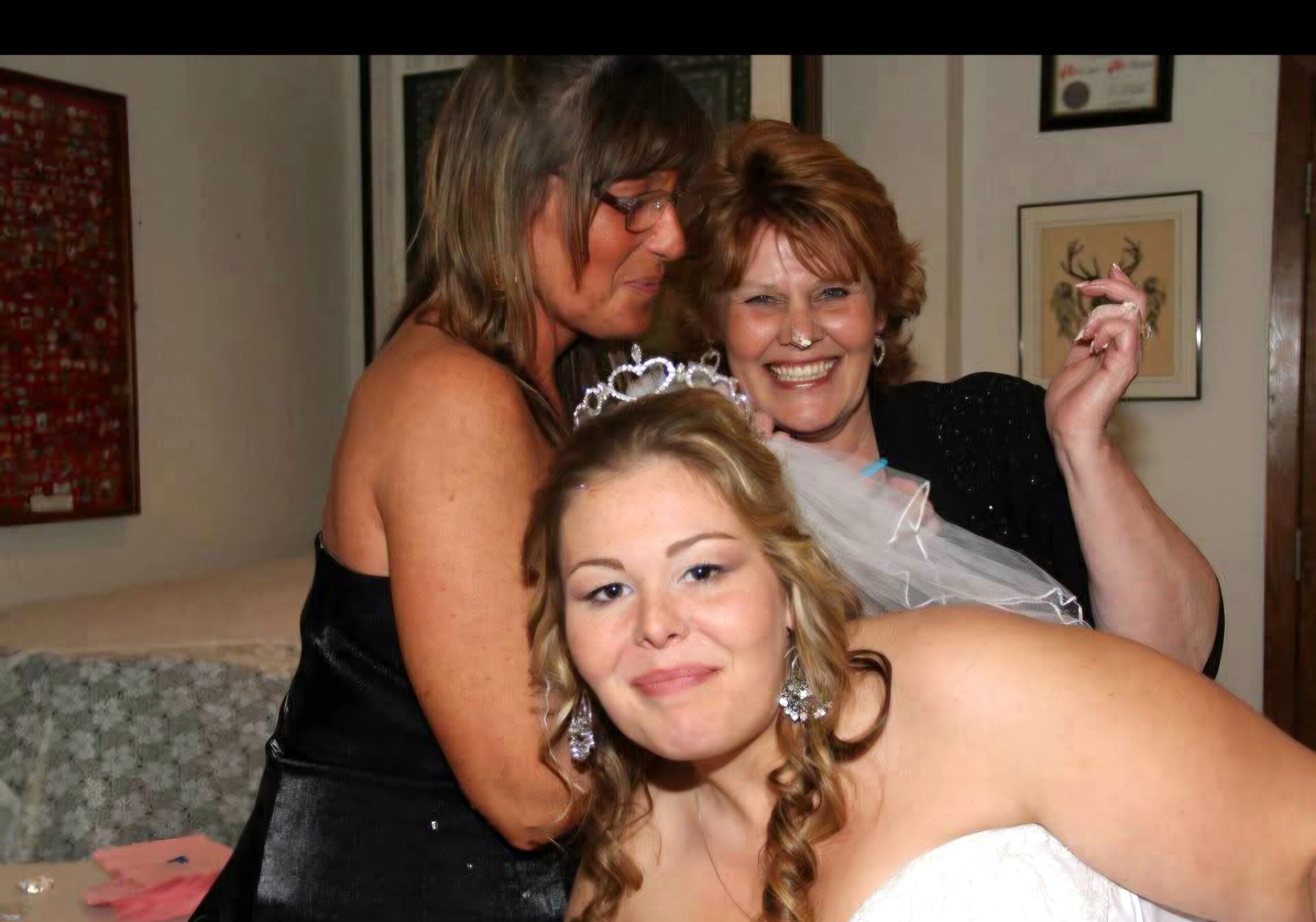 Smiling bride in a white dress with veil, posing with two friends at a celebration in a home setting.