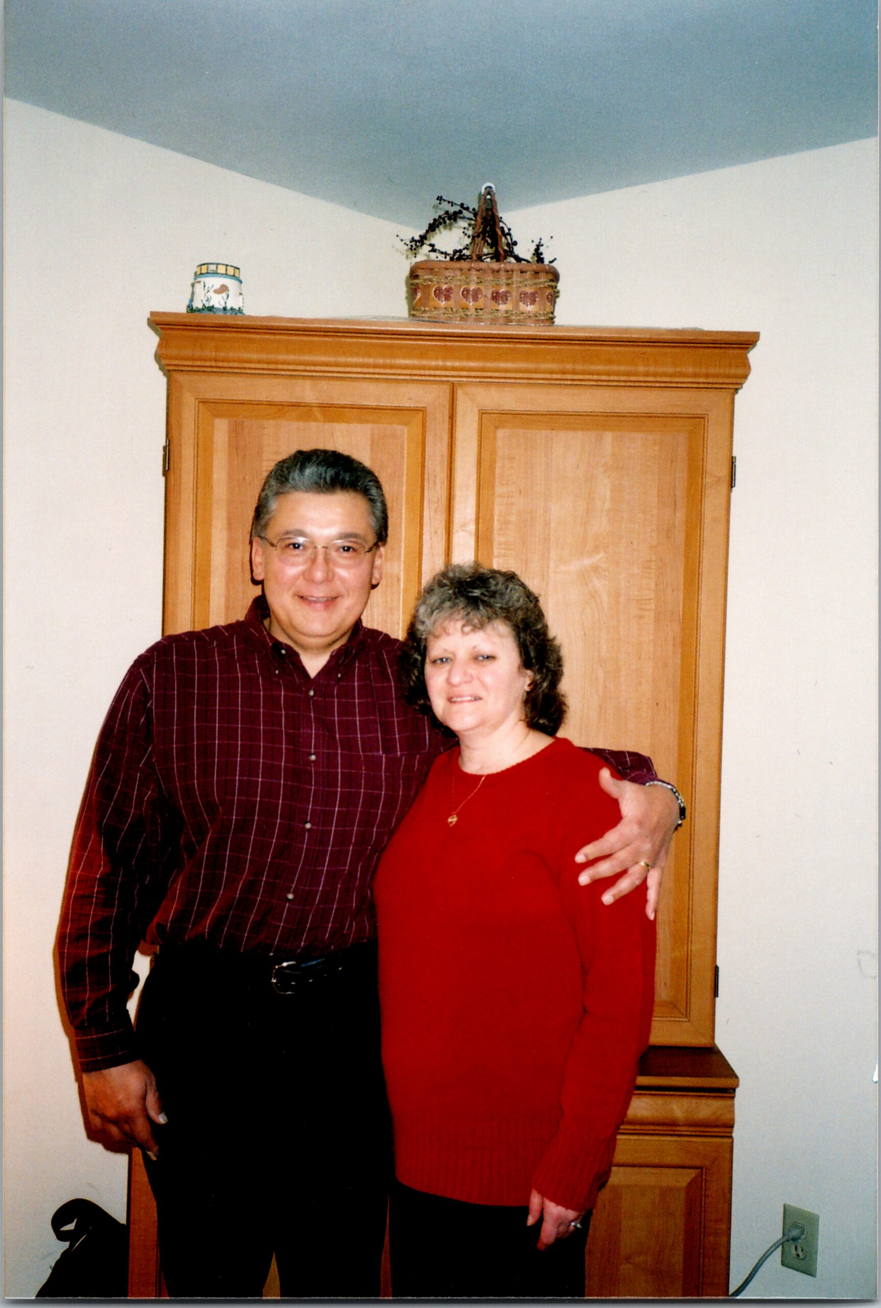 Smiling couple standing in front of a tall wooden cabinet indoors, with a wicker basket and ceramic jar on top shelf behind them.