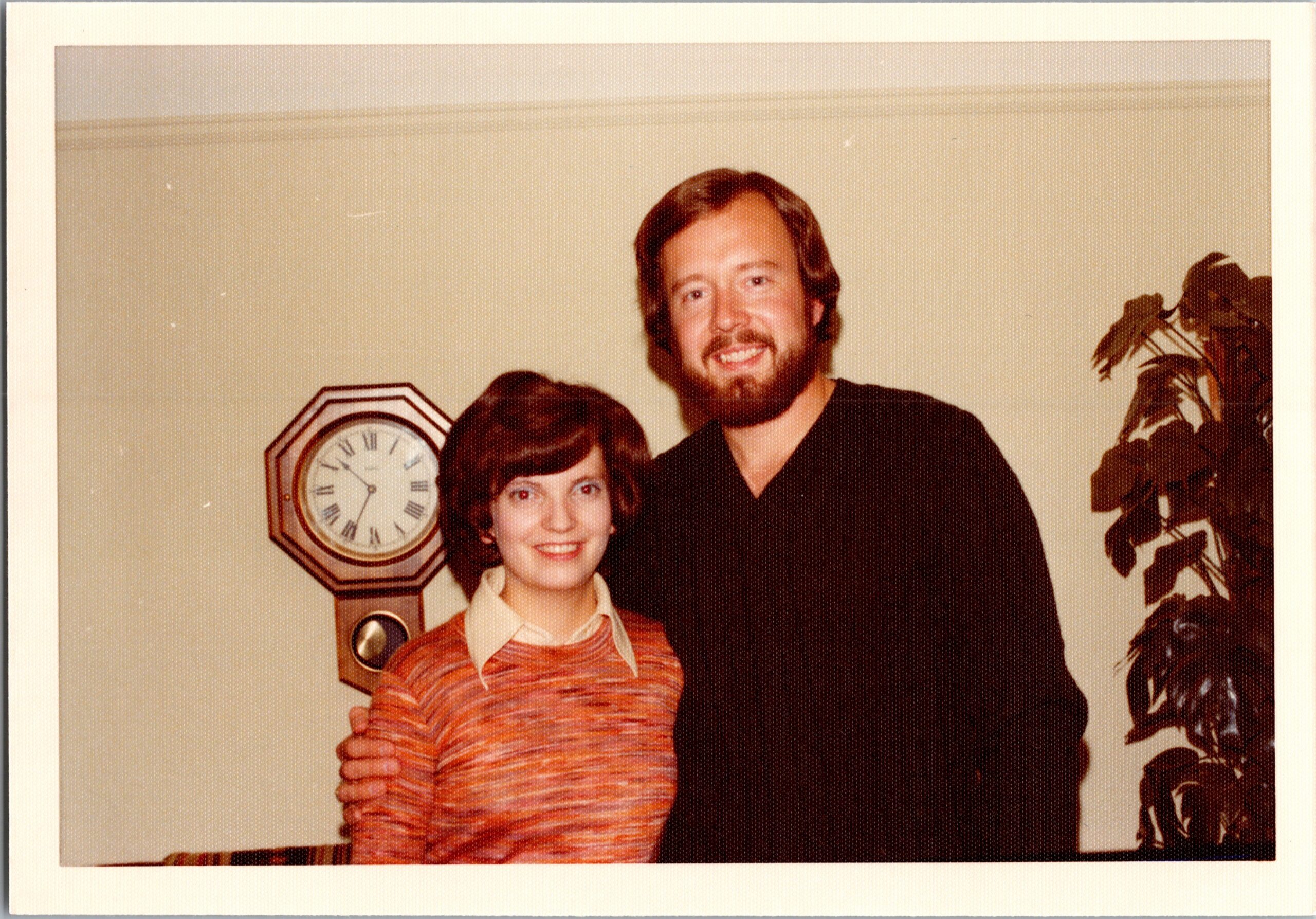 Smiling couple posing indoors: woman in orange striped sweater in front, man with beard behind her, with an octagonal wall clock visible to the left and a plant on the right edge.