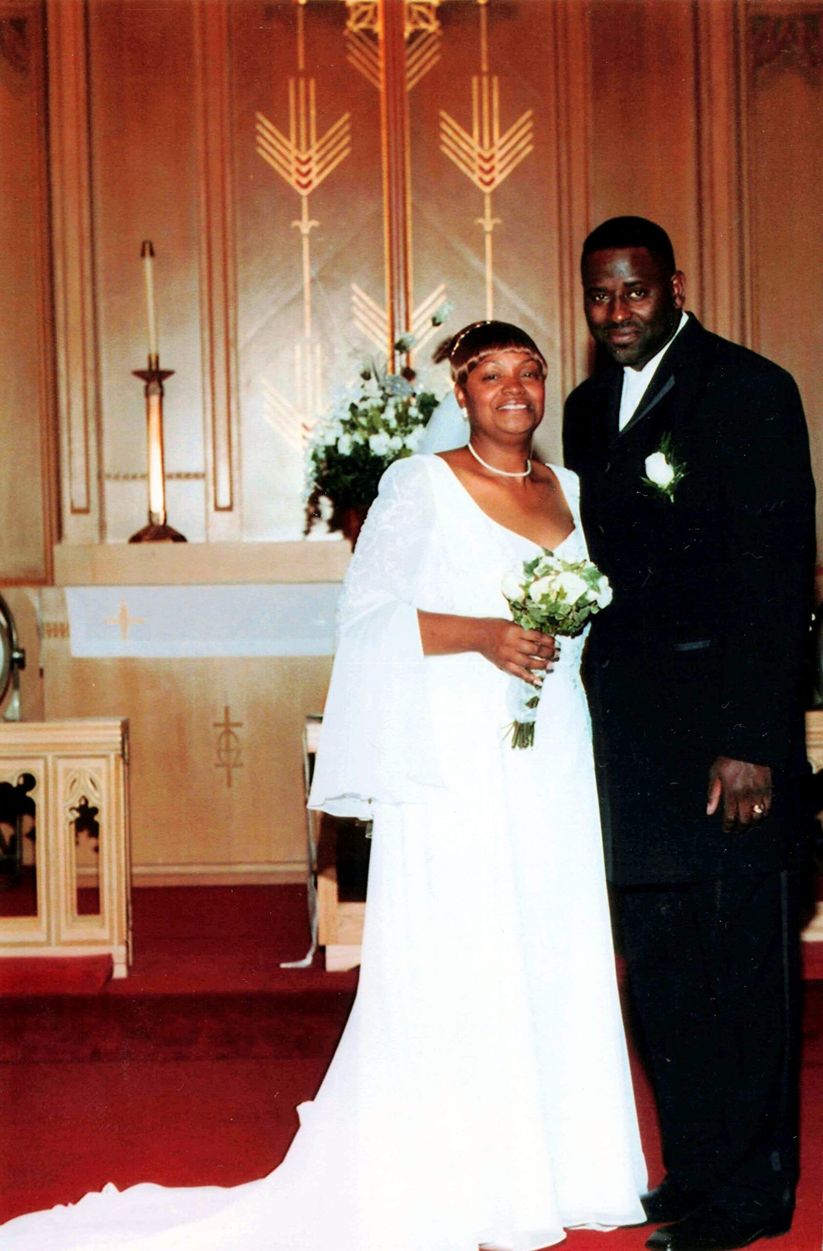 Bride in a white wedding dress and veil holds a bouquet beside her groom in a black suit inside a church altar setting with candles and flowers behind them.