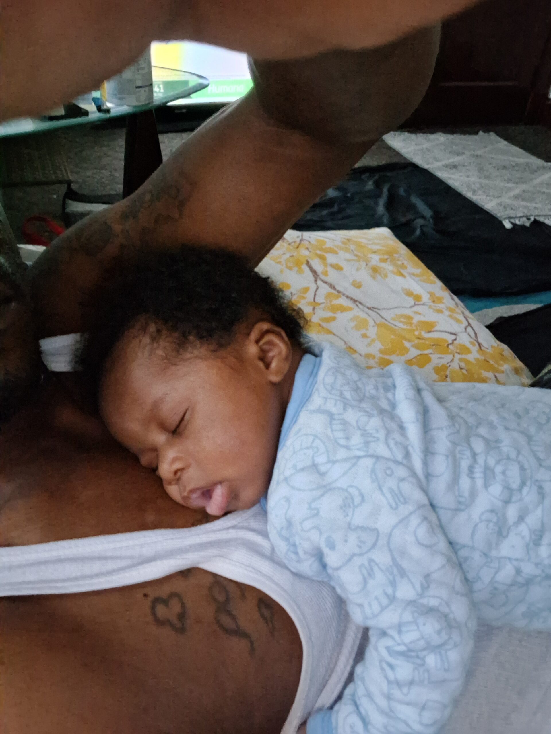 Baby sleeps cuddled against an adult's chest, arm raised above the head; the baby is in a blue onesie and lies on a floral pillow cover.
