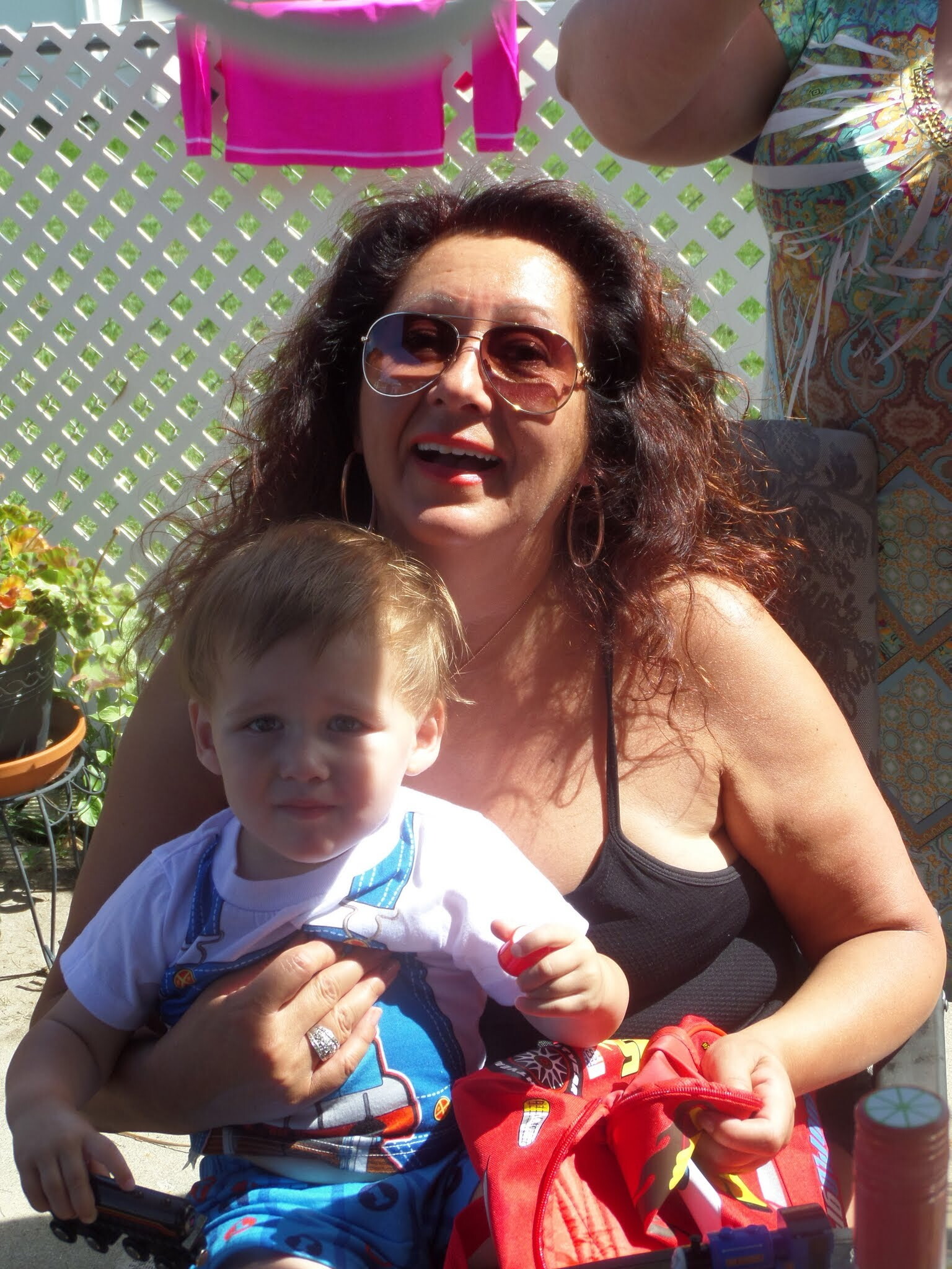 Woman with sunglasses holding a smiling baby outdoors, with a lattice fence behind them for support or privacy.