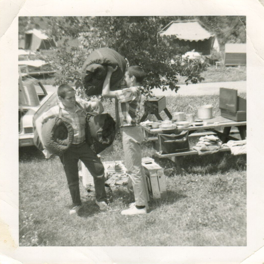 Two men loading camping gear at a park: one passes a rolled sleeping pad to the other, with a car, picnic table, and supplies in the background.