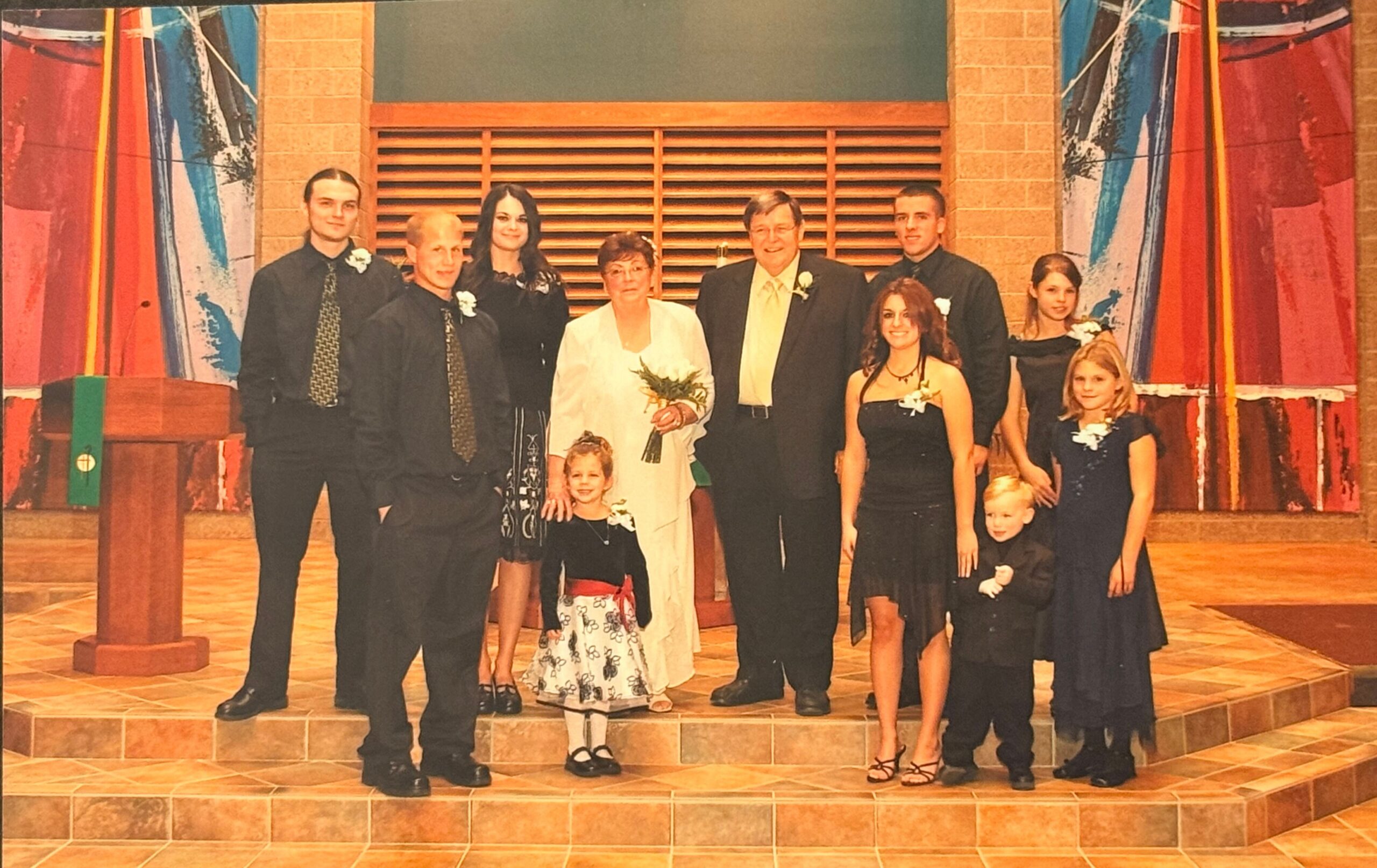 Group portrait on a stage with a bride and groom in the center, flanked by adults and children in formal black attire, holding flowers.