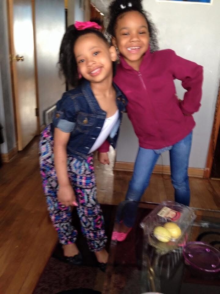 Two smiling young girls posing indoors together, one in a denim jacket and patterned pants and the other in a magenta hoodie.