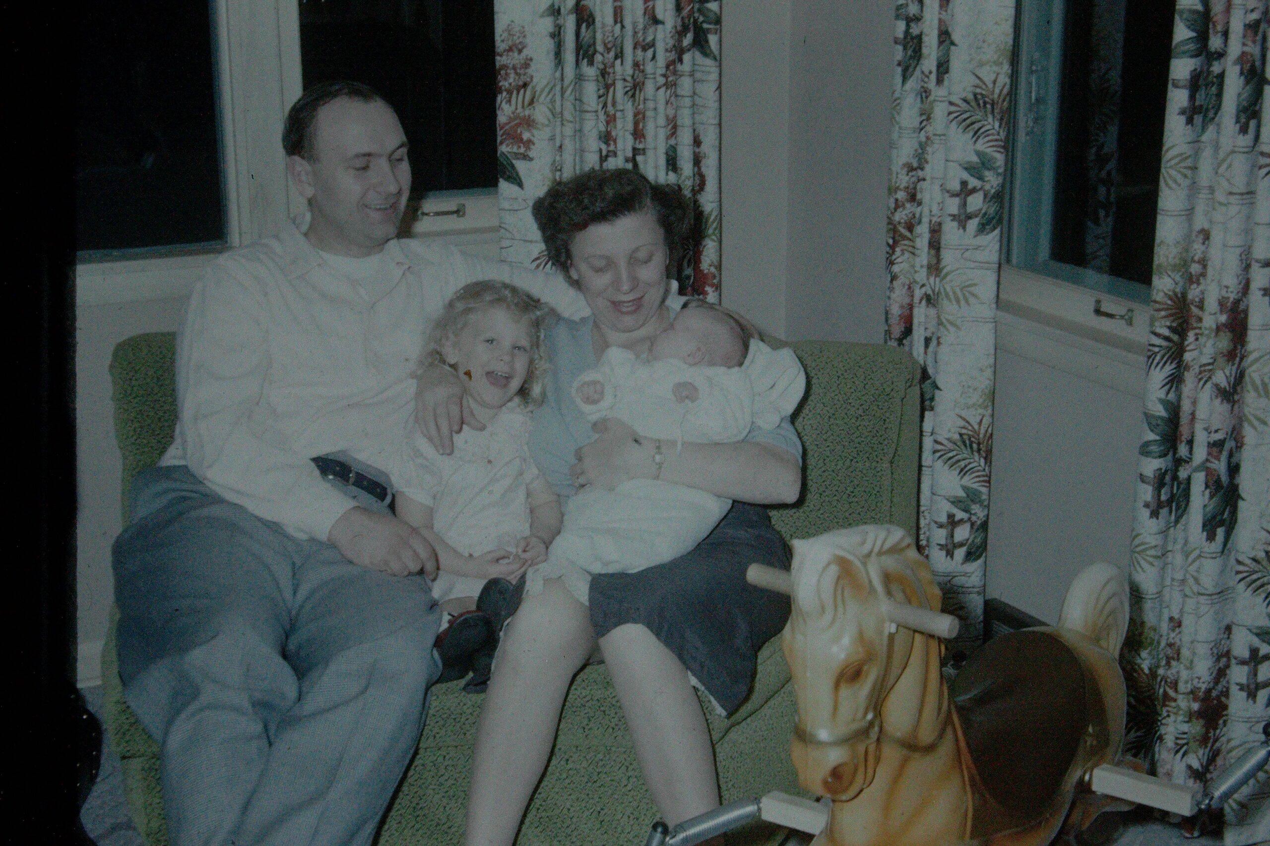 Man and woman sit on a green couch with two young children: a baby in the woman’s arms and a smiling toddler beside her, in a living room with patterned curtains.