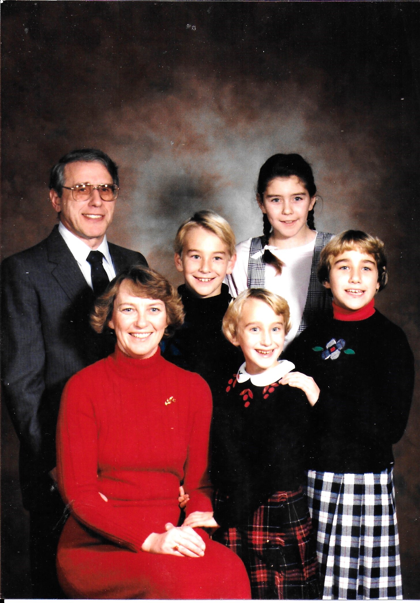 Studio family portrait: an older man in a suit and tie on the left, a smiling woman in a red sweater seated, and several children standing and posing in colorful outfits behind and beside them.
