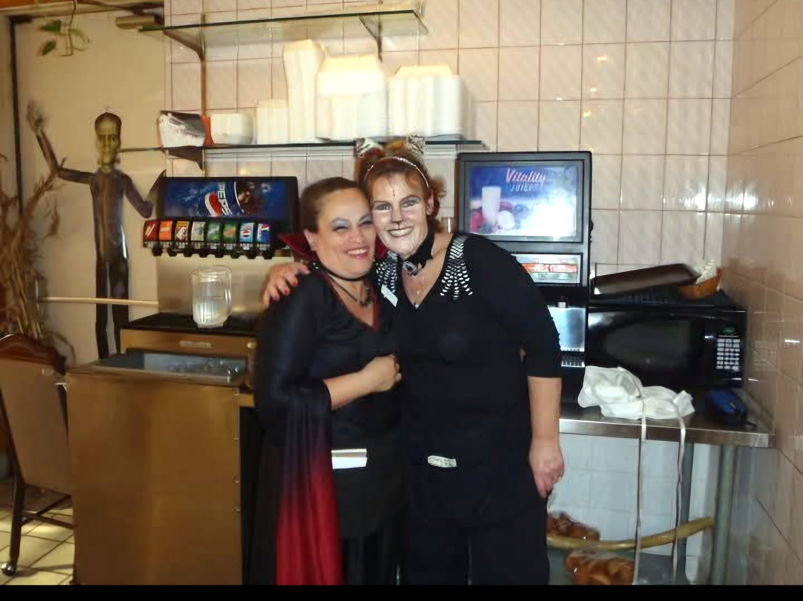 Two women in Halloween costumes posing and smiling together behind a counter, with a skeleton figure in a display area nearby.