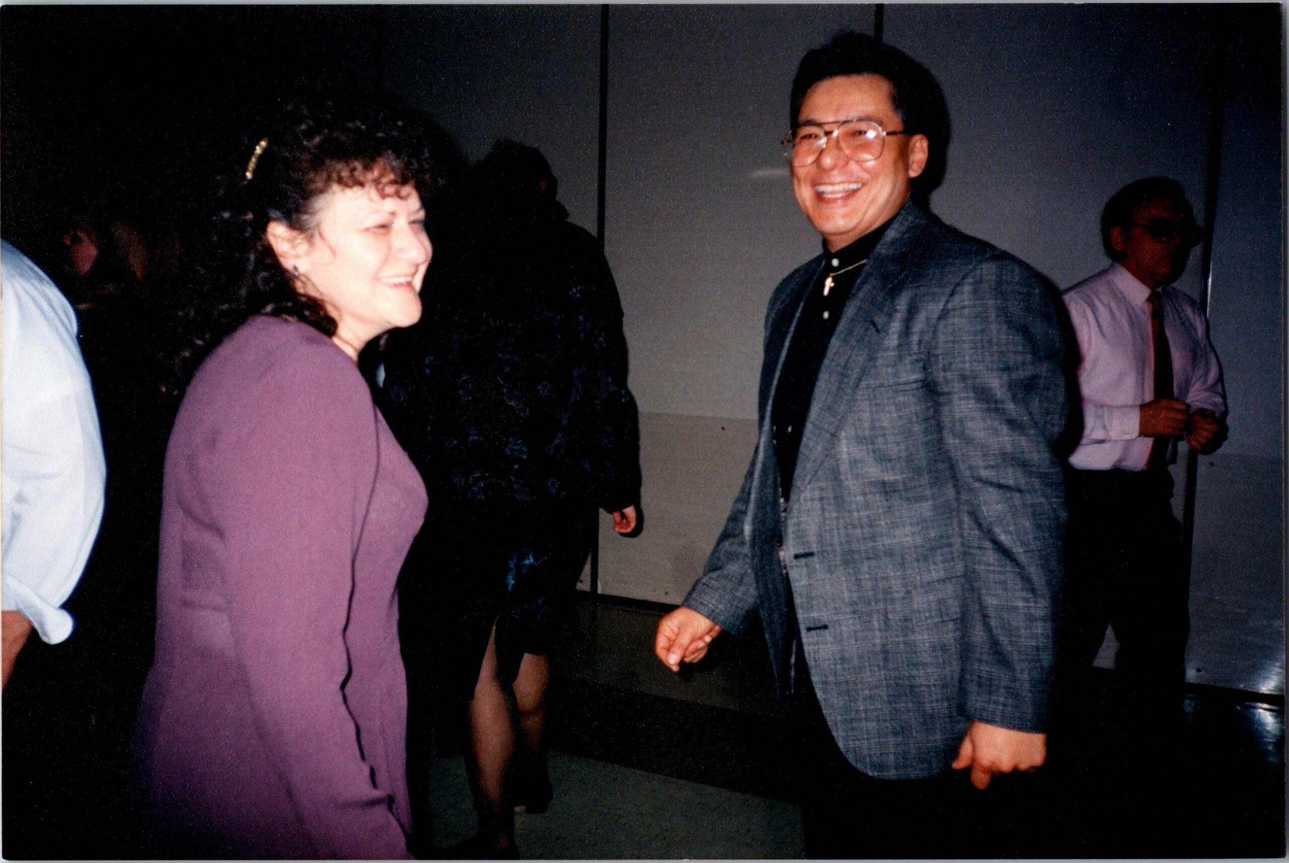 Two adults at a social event share a smile and converse, with a woman in a purple dress on the left and a man in a gray plaid suit on the right.