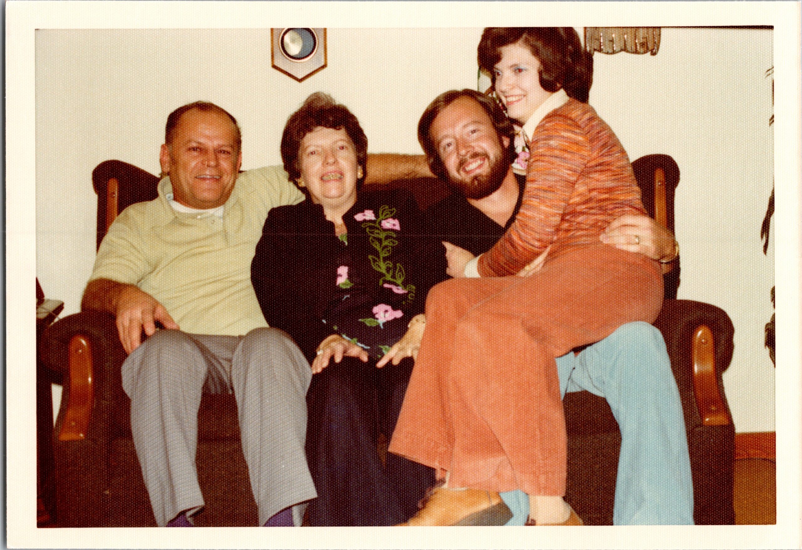 Four adults sit closely on a sofa, smiling for a casual group photo in a living room.