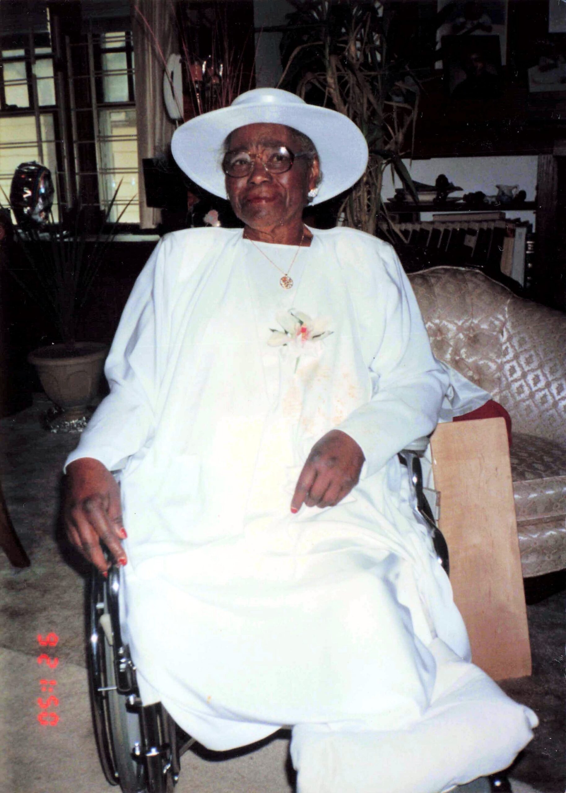 Older woman in a light blue dress and wide white hat sits in a wheelchair indoors, with a flower corsage on her chest.