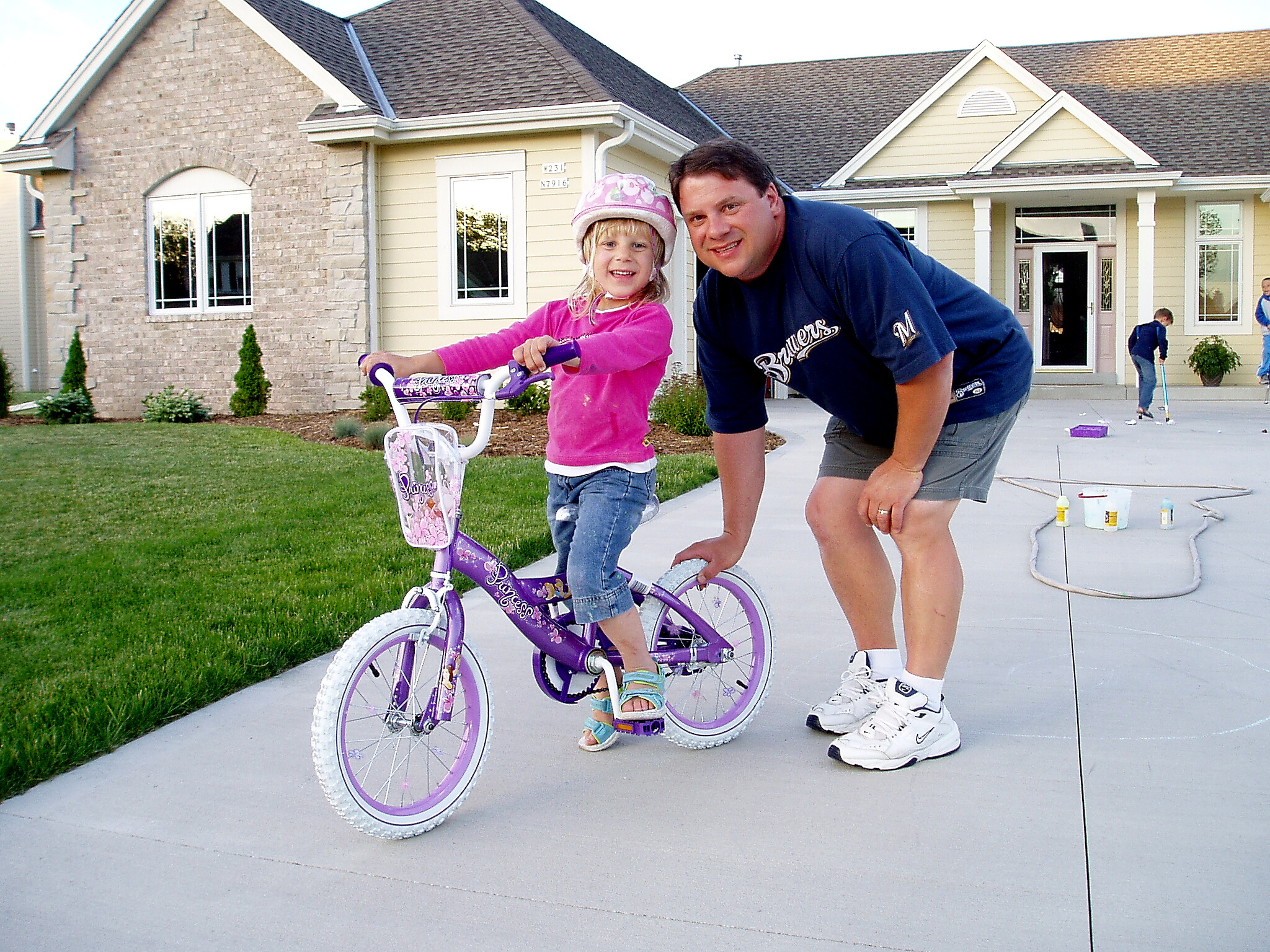 A girl rides a purple bicycle with training wheels while an adult man leans beside her in a suburban driveway.