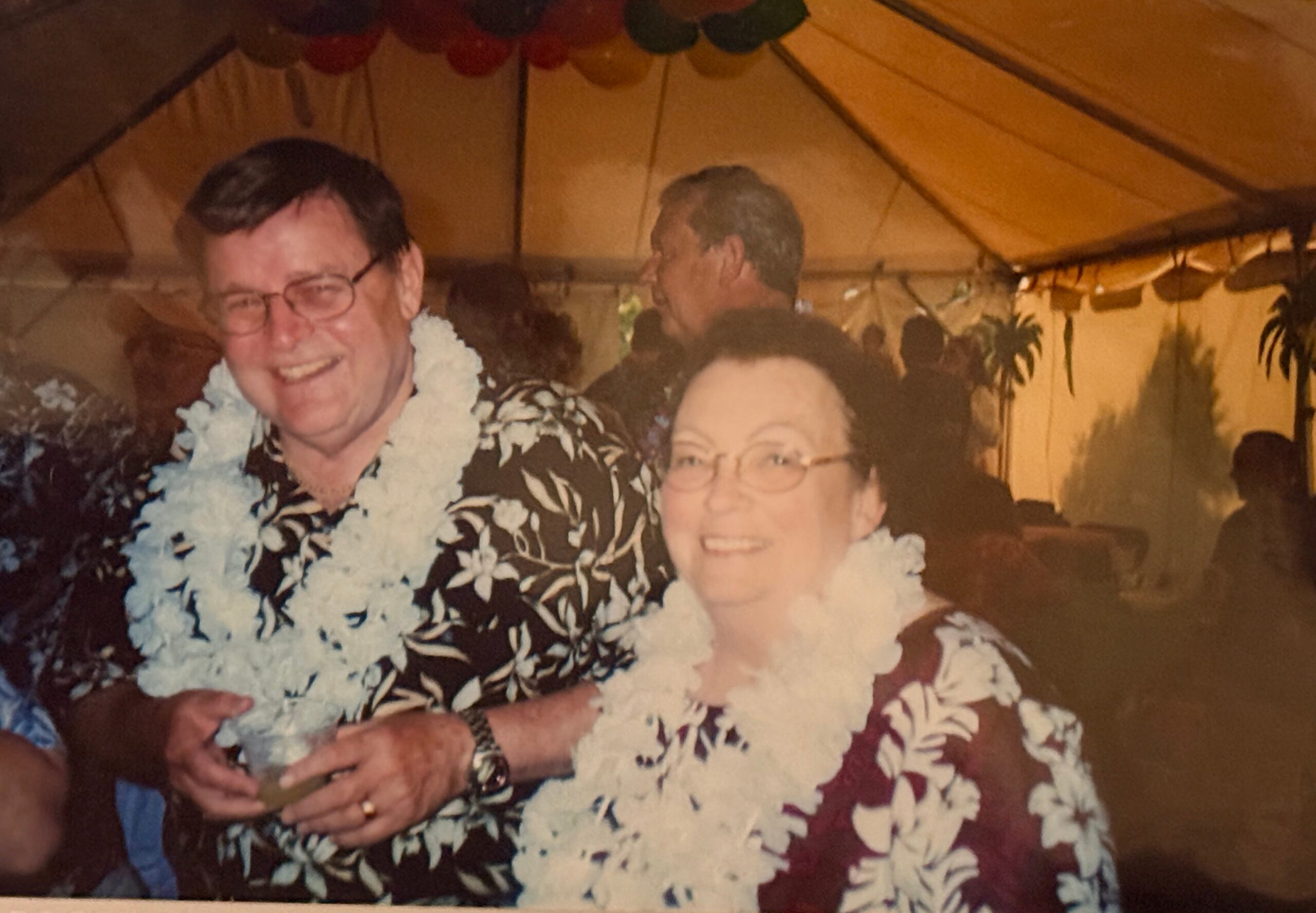 Smiling couple in Hawaiian shirts wearing flower leis at a tropical-themed party indoors.