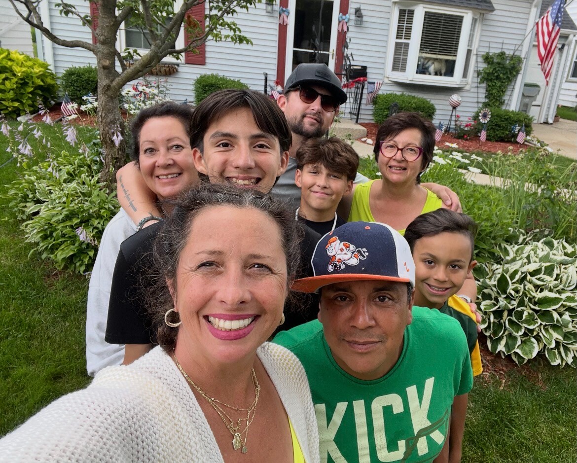 Smiling group of eight posing for a selfie on the front lawn of a suburban home with American flags and garden plants behind them.