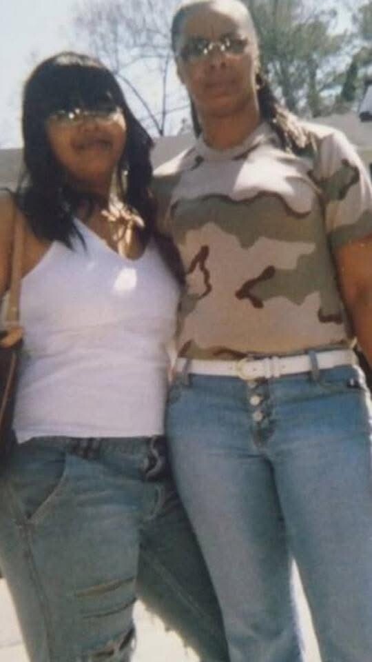 Two women posing outdoors in daylight; one wearing a white tank top and jeans, the other in a camouflage shirt, jeans, and sunglasses.