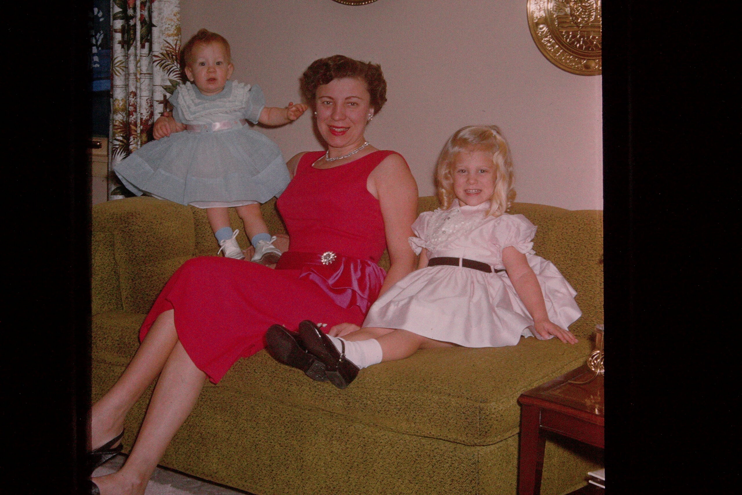 Mother in a red dress sits on a green sofa with two young girls, one on her arm in a blue dress and the other in a white dress beside her on the couch, all smiling.