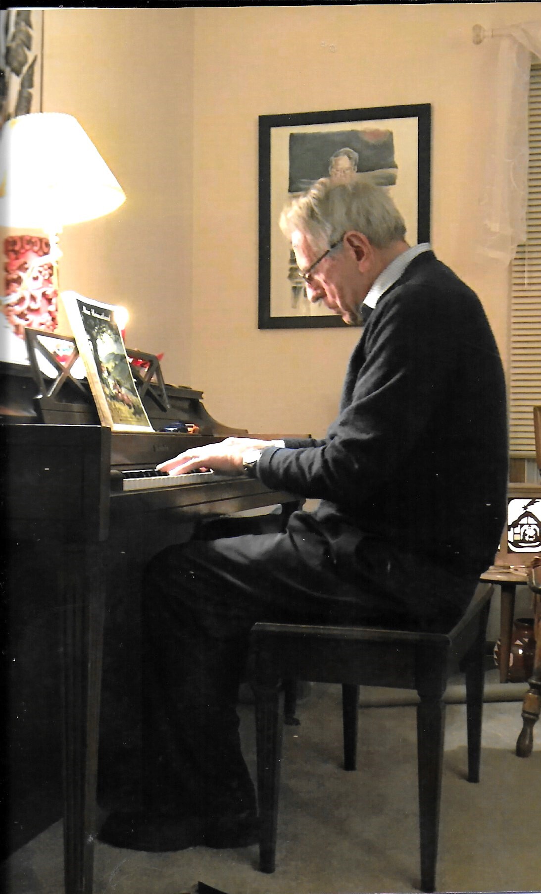 Elderly man in a dark sweater playing a upright piano in a living room, with a lamp and framed artwork behind him.