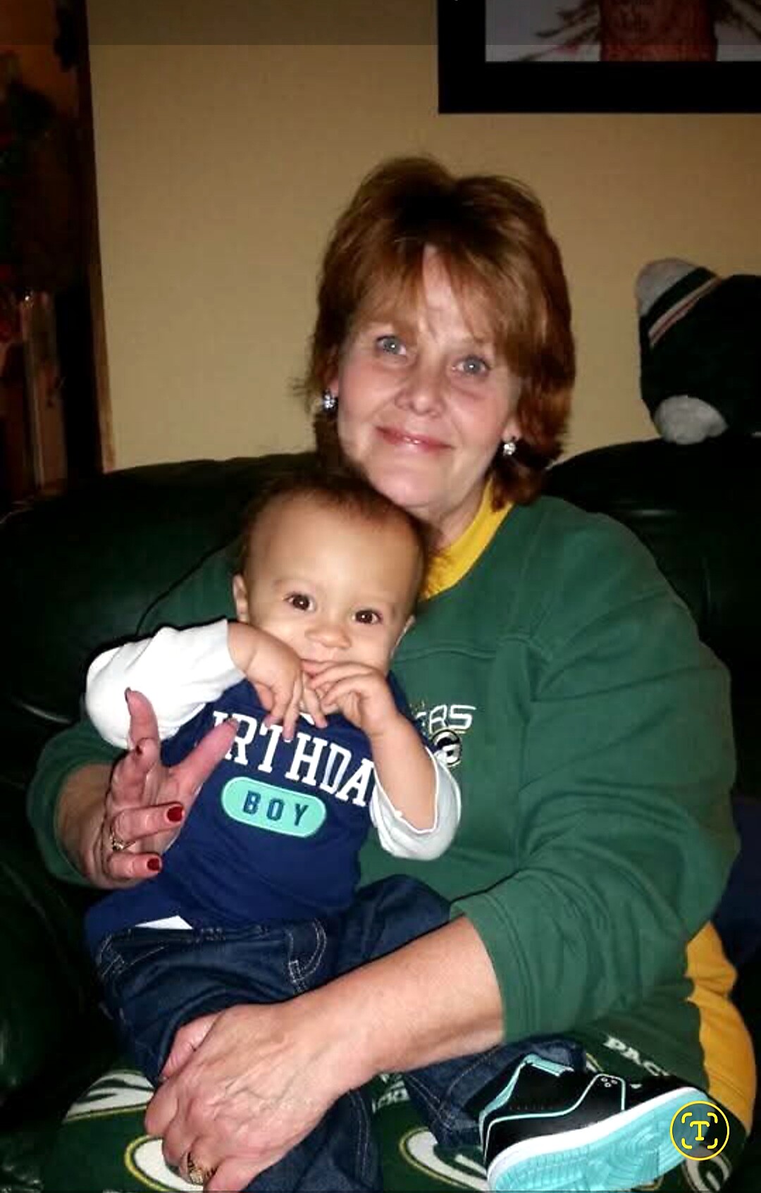 Elderly woman with short brown hair sits on a couch, holding a smiling baby boy in a blue 'Birthday Boy' outfit.