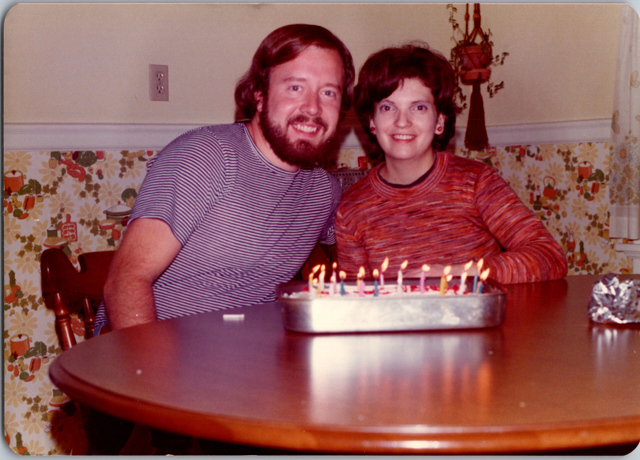 Smiling couple sits at a table with a birthday cake and lit candles in front of them.