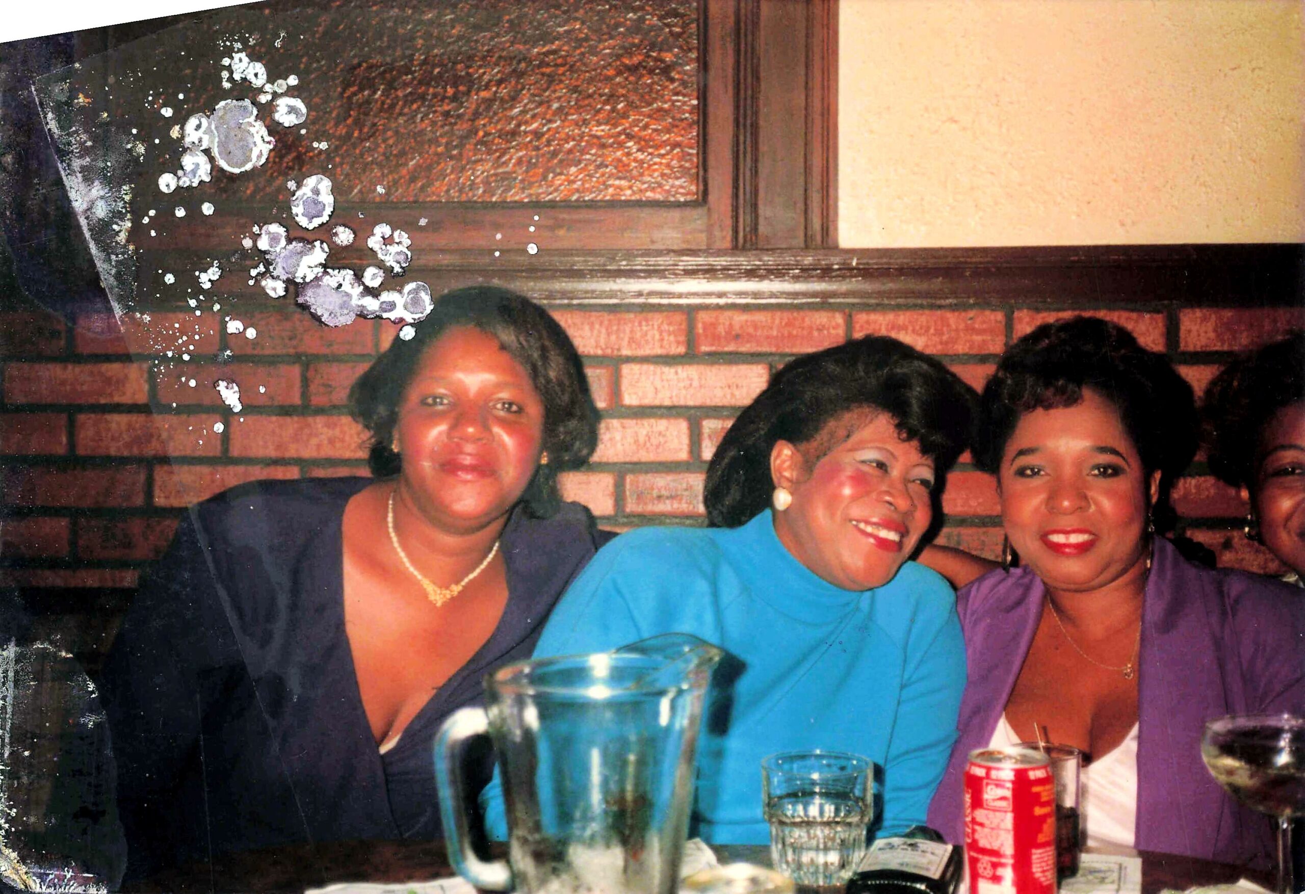 Group of smiling women around a table at a social gathering, brick wall background; one drinks glass in foreground; vintage photo with paint splatter marks on left