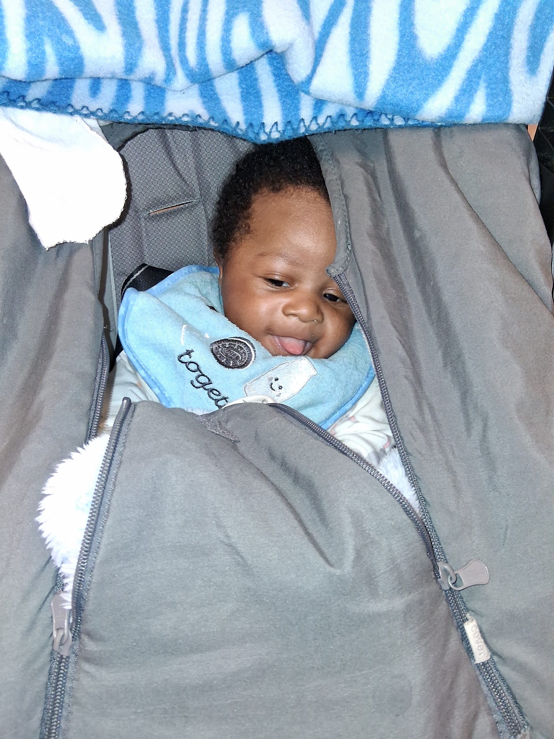 Smiling infant bundled in a gray jacket in a car seat with a blue blanket overhead.