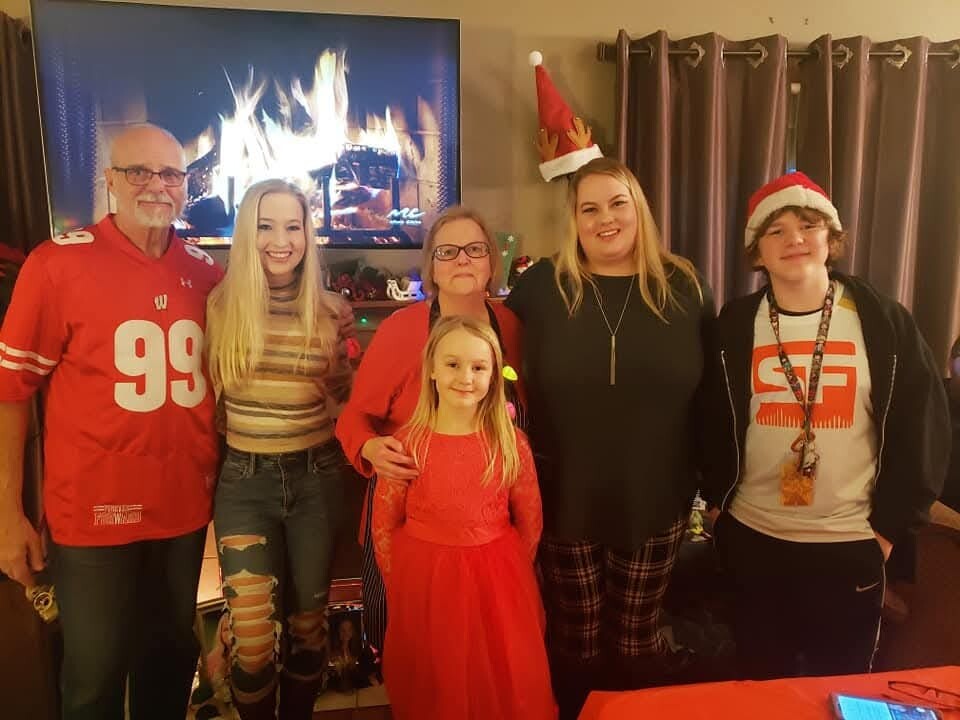 Group of six people posing in a cozy living room with a lit fireplace, Christmas hats, and festive outfits (one in a red football jersey).