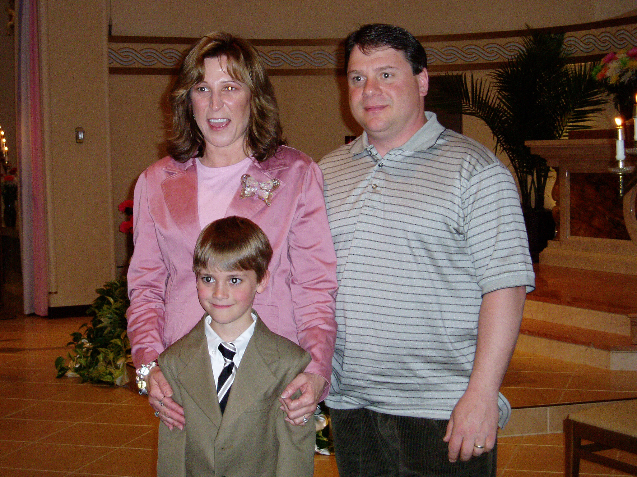 Woman in pink blouse beside a man in a striped polo; a boy in a suit stands in front, indoors. (Informative group portrait)