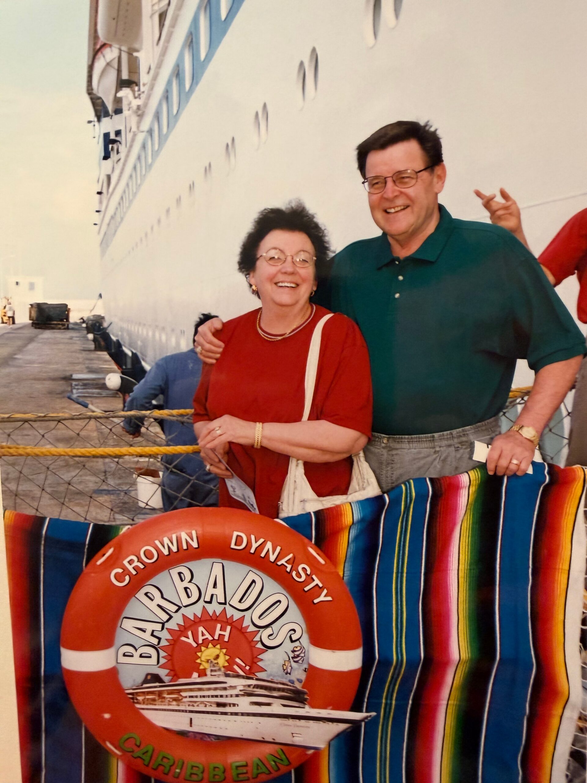 Smiling older couple posing in front of a large cruise ship, with a red Barbados life ring and colorful striped fabric nearby.