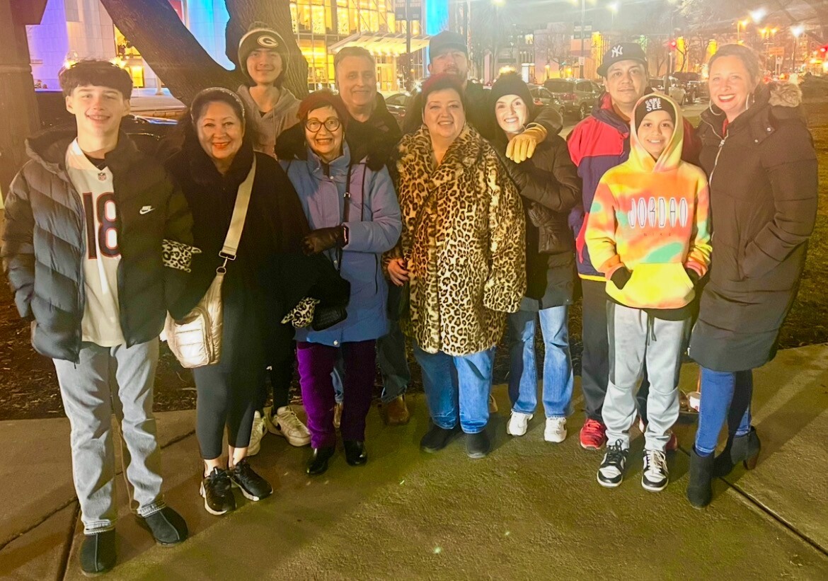 A diverse group of about 12 people in winter coats posing together on a city sidewalk at night, smiling for the camera.