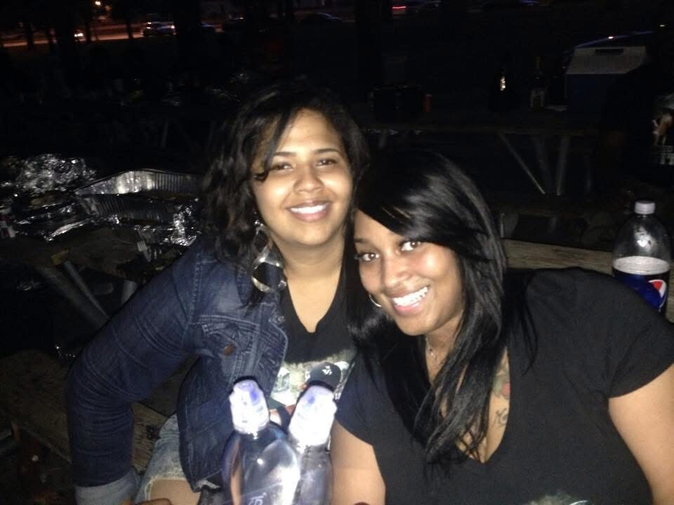 Two smiling women posing closely at night, leaning together at a picnic table with bottled drinks nearby.