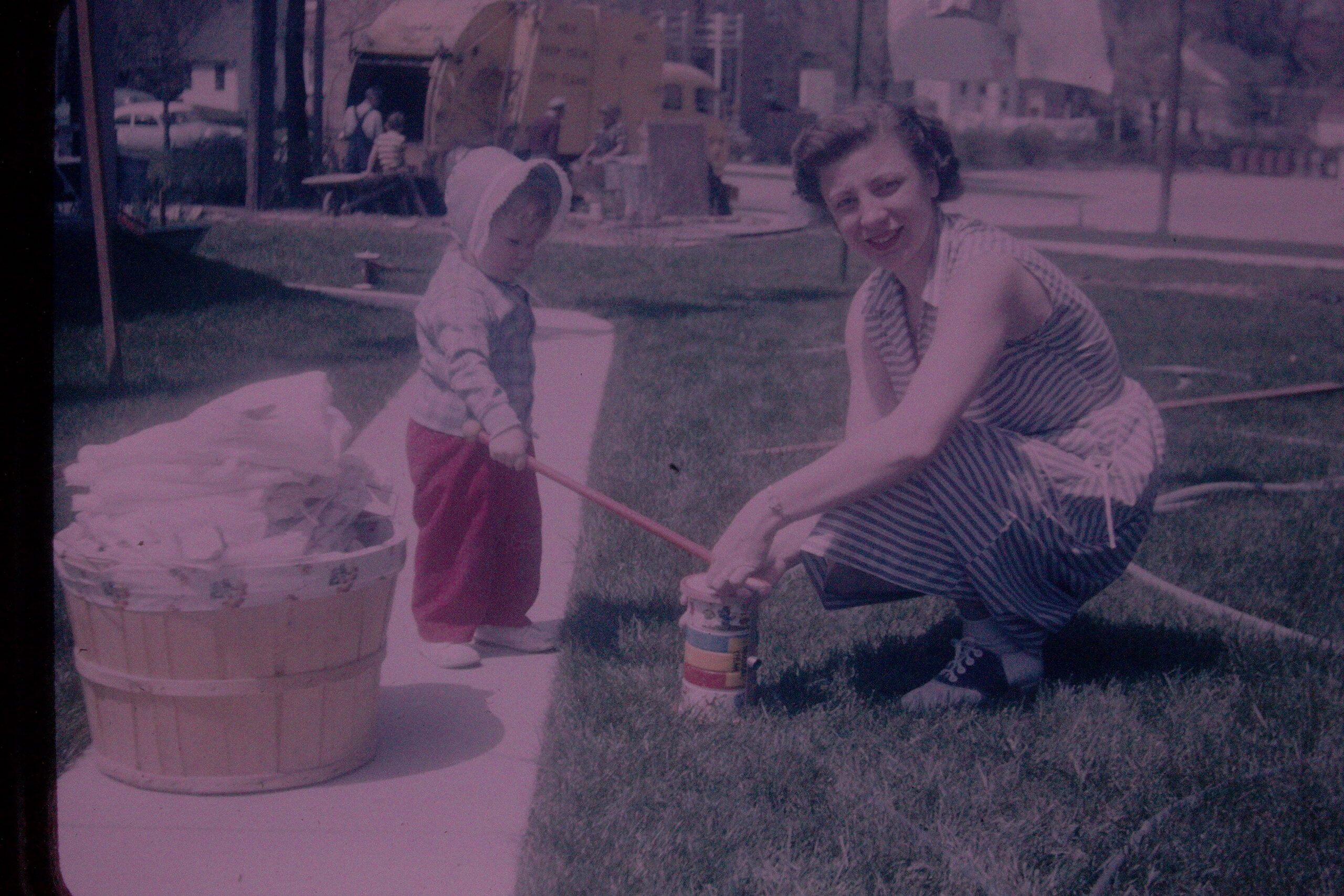 A woman in a striped dress kneels on grass beside a young child in a hooded jacket, as the child uses a stick near a large wooden barrel planter outdoors.