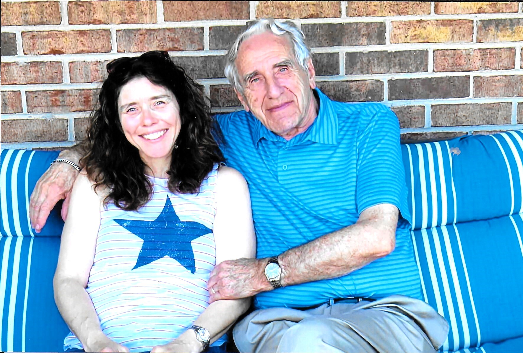 Smiling older man and woman sitting close together on a blue striped outdoor couch against a brick wall.