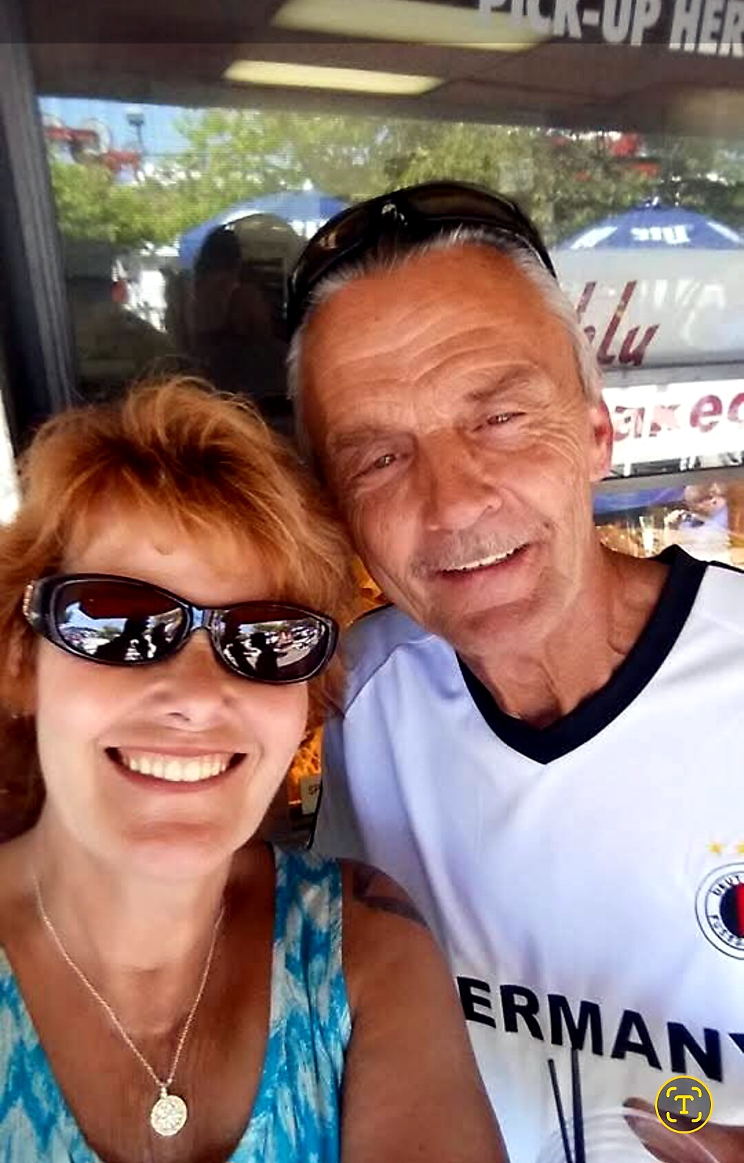 Smiling woman and man taking a close selfie outdoors, woman wearing sunglasses and a turquoise top, man with gray hair and a white shirt with dark trim