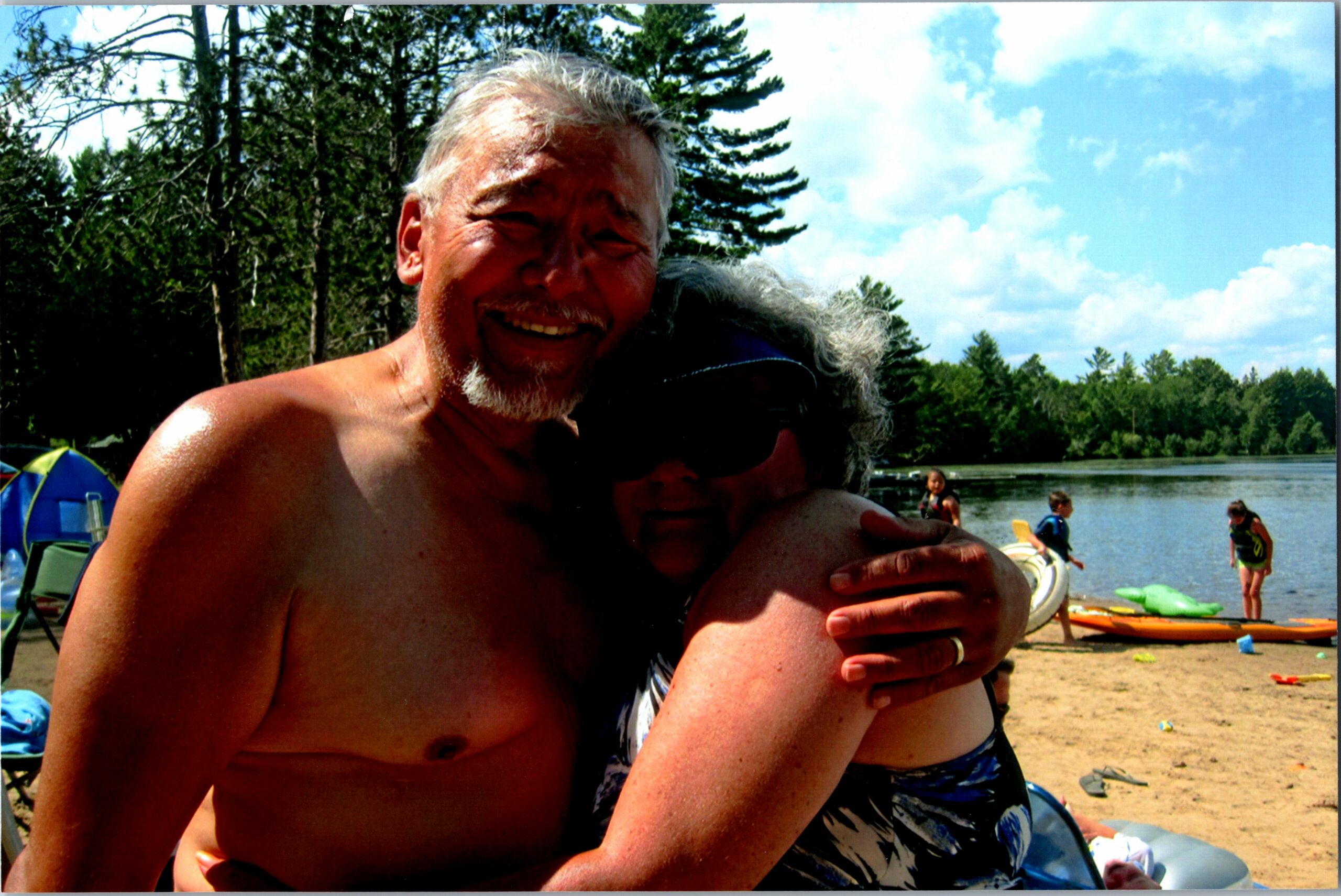Two adults hug on a sandy lakeshore with trees and kayaks nearby on a sunny day by the lake.