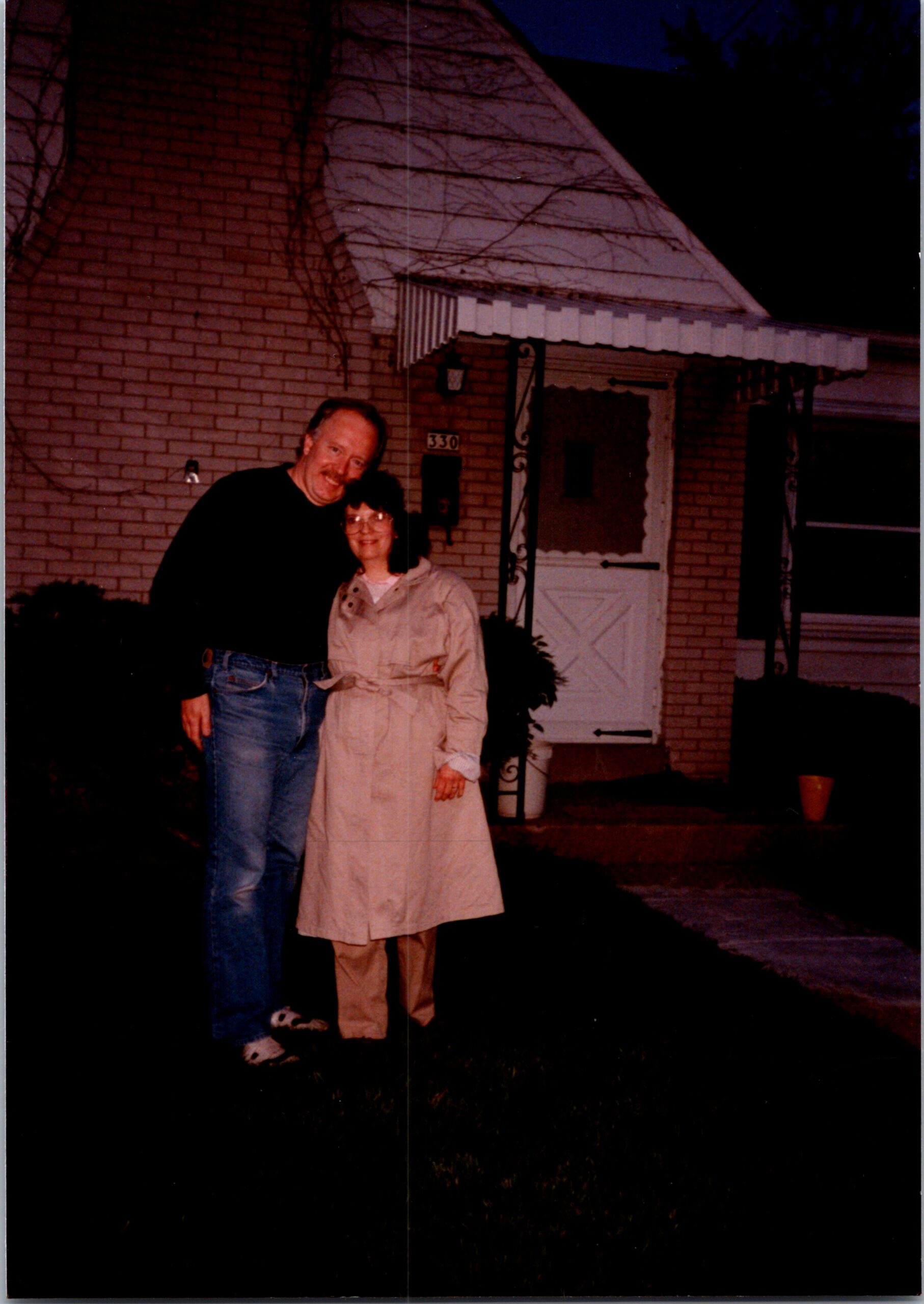 A man and a woman stand together outside a brick house at night, smiling for the photo in front of the entrance labeled 330.