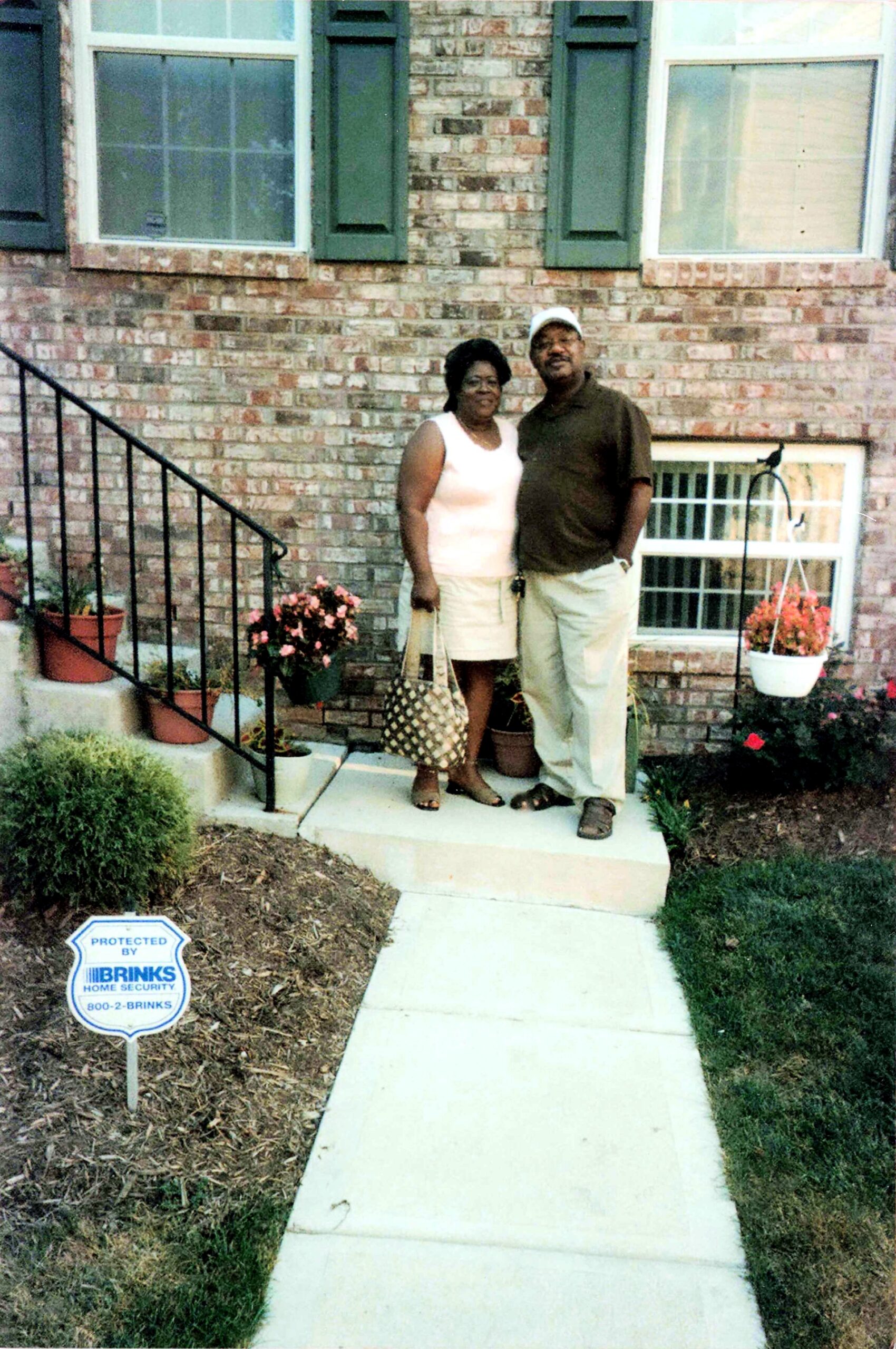 African American couple standing together on a concrete front walkway in front of a brick house with green shutters, potted flowers, and a Brinks home security sign in the yard.