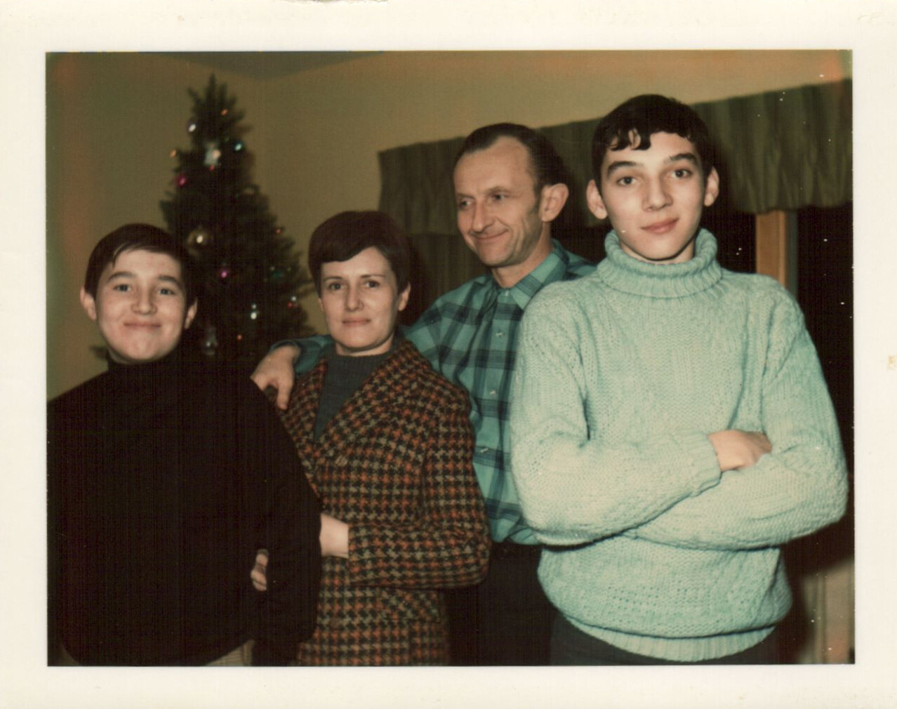 Four family members pose for a living-room photo with a Christmas tree in the background, a mother, father, and two sons smiling.
