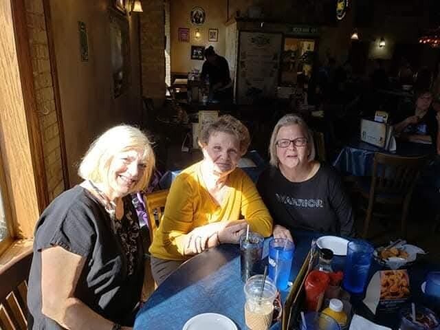 Three friends pose for a photo at a cozy restaurant table, smiling with drinks and snacks in view. (Sunlight on the left)