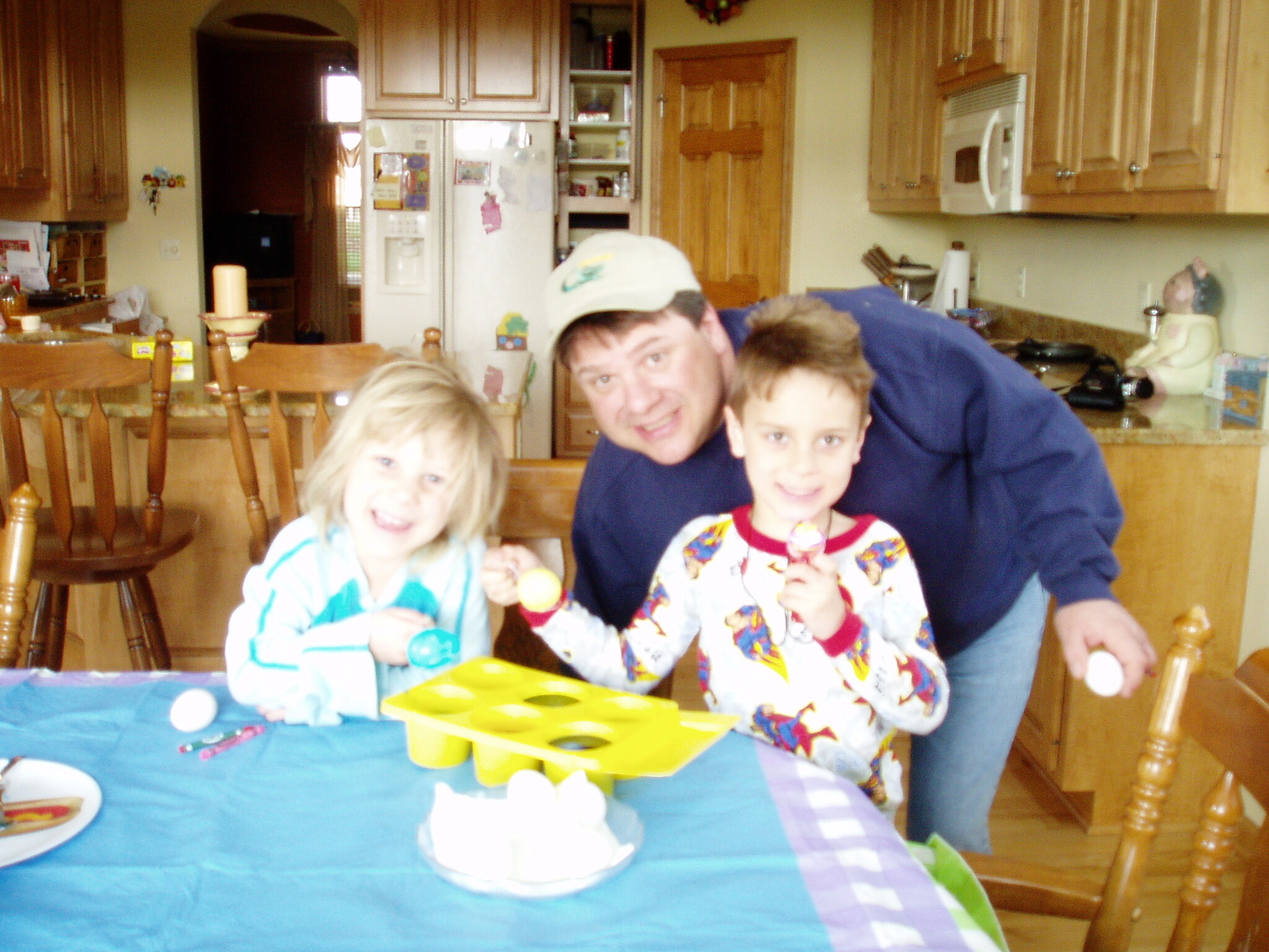 A man hugs two smiling children at a kitchen birthday party, with a blue tablecloth and snack items on the table in front.