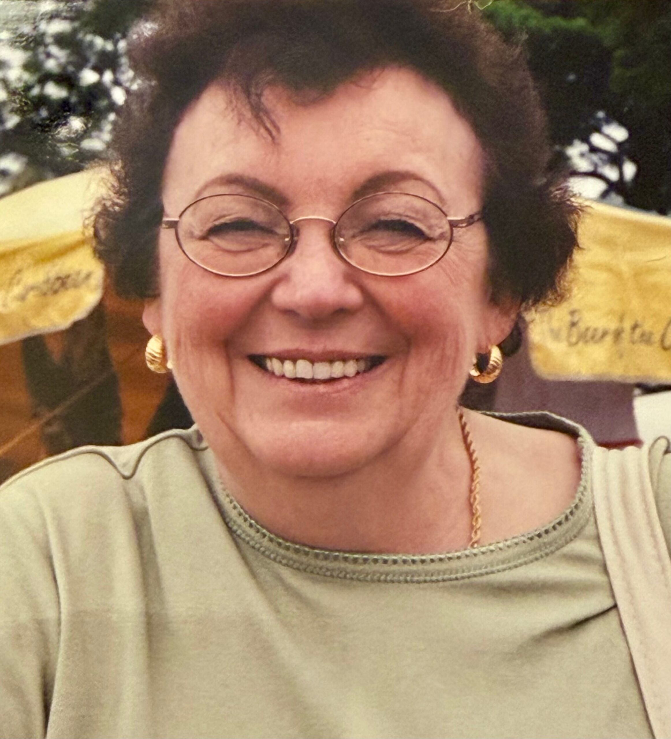 Close-up of a smiling woman with short dark hair, round glasses, and gold hoop earrings, wearing a light-colored top.