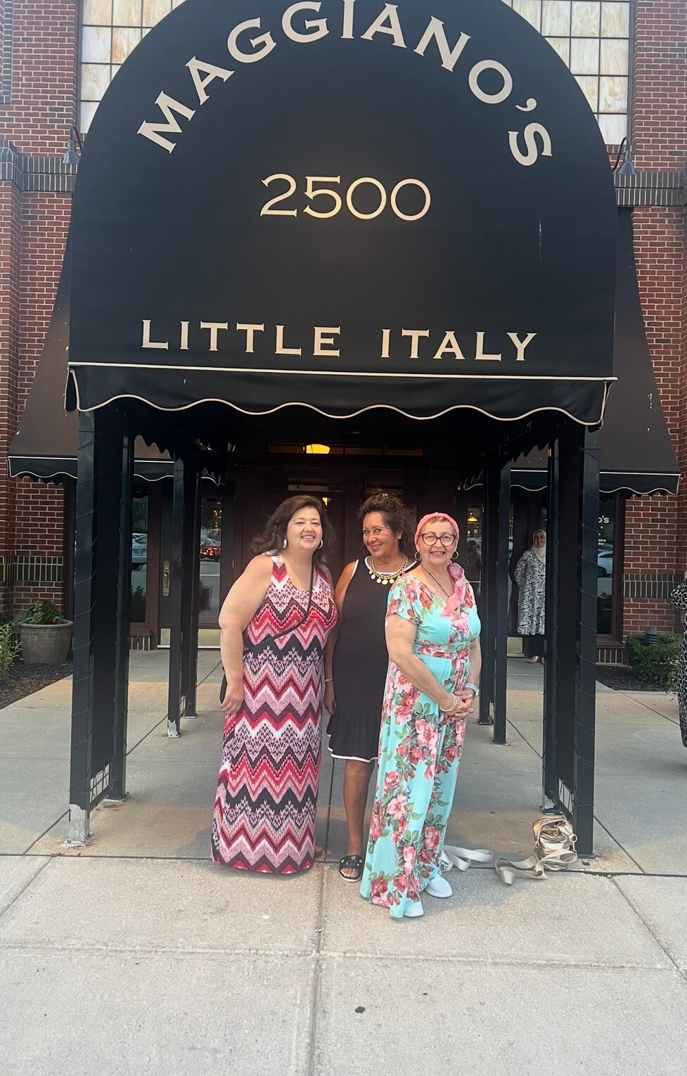 Three women pose together outside Maggiano's Little Italy restaurant (address 2500) in front of the black awning.