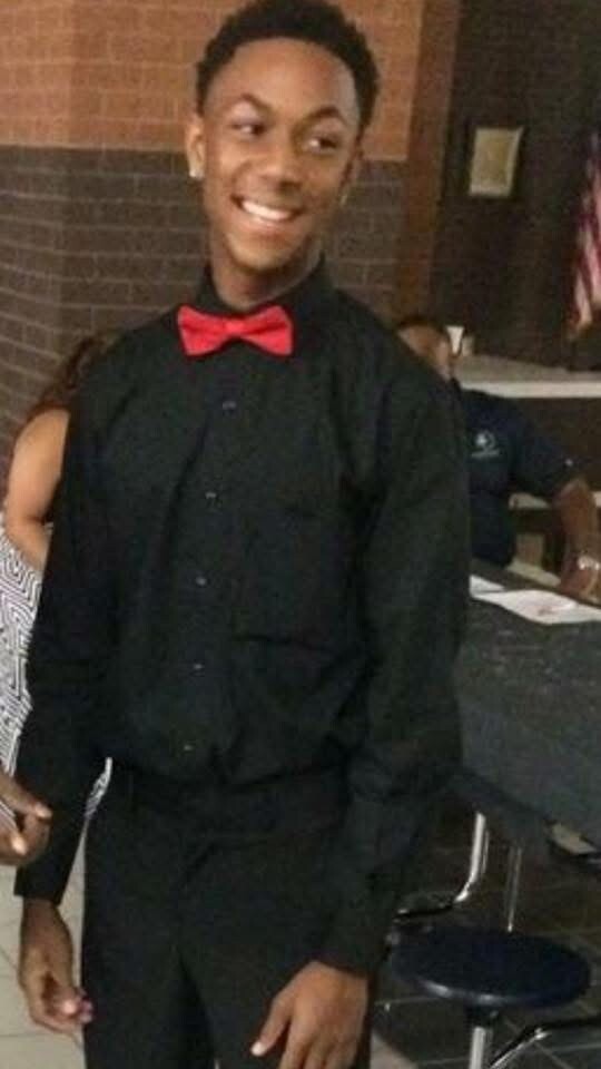 Smiling young man in a black shirt with a red bow tie at an indoor event, brick wall behind him.