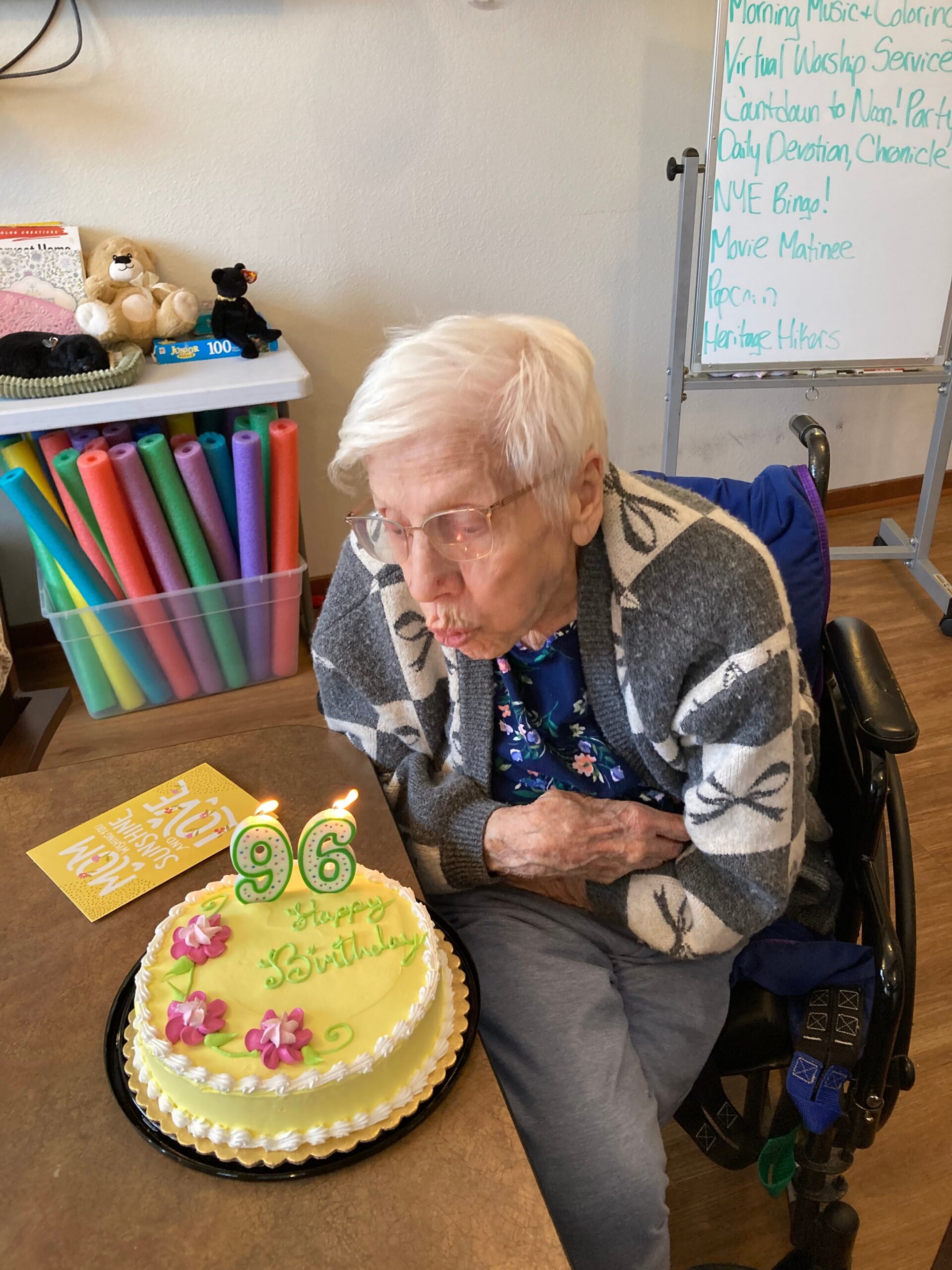 Elderly woman in a wheelchair leans toward a yellow birthday cake with lit '96' candles; colorful décor and stuffed animals on a shelf in the background.
