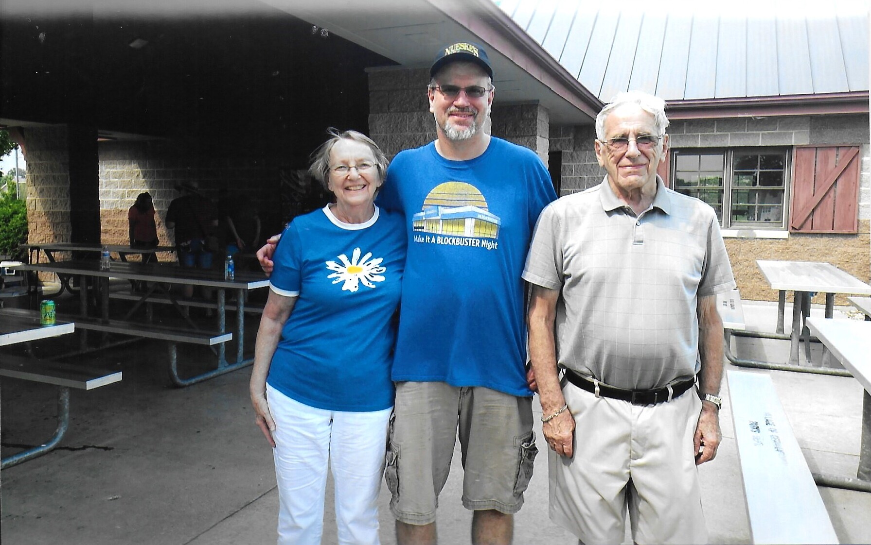 Three adults pose together outside a pavilion, smiling at camera; man in the middle wears a blue shirt and cap, flanked by a woman in a blue daisy shirt and an older man in light polo.
