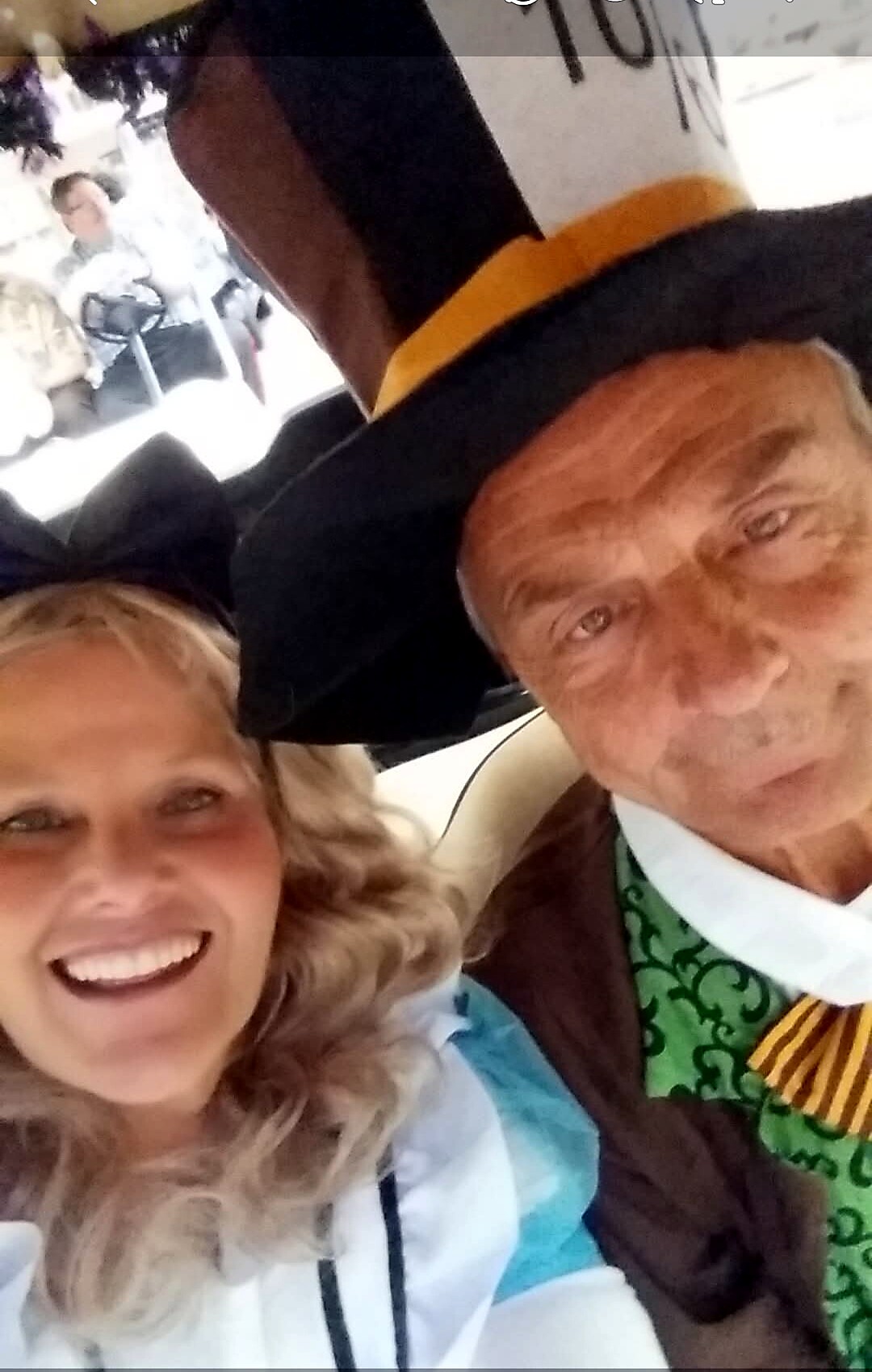 Smiling woman and an older man take a selfie at a graduation ceremony; the man wears a cap and a gown with green and yellow trim.