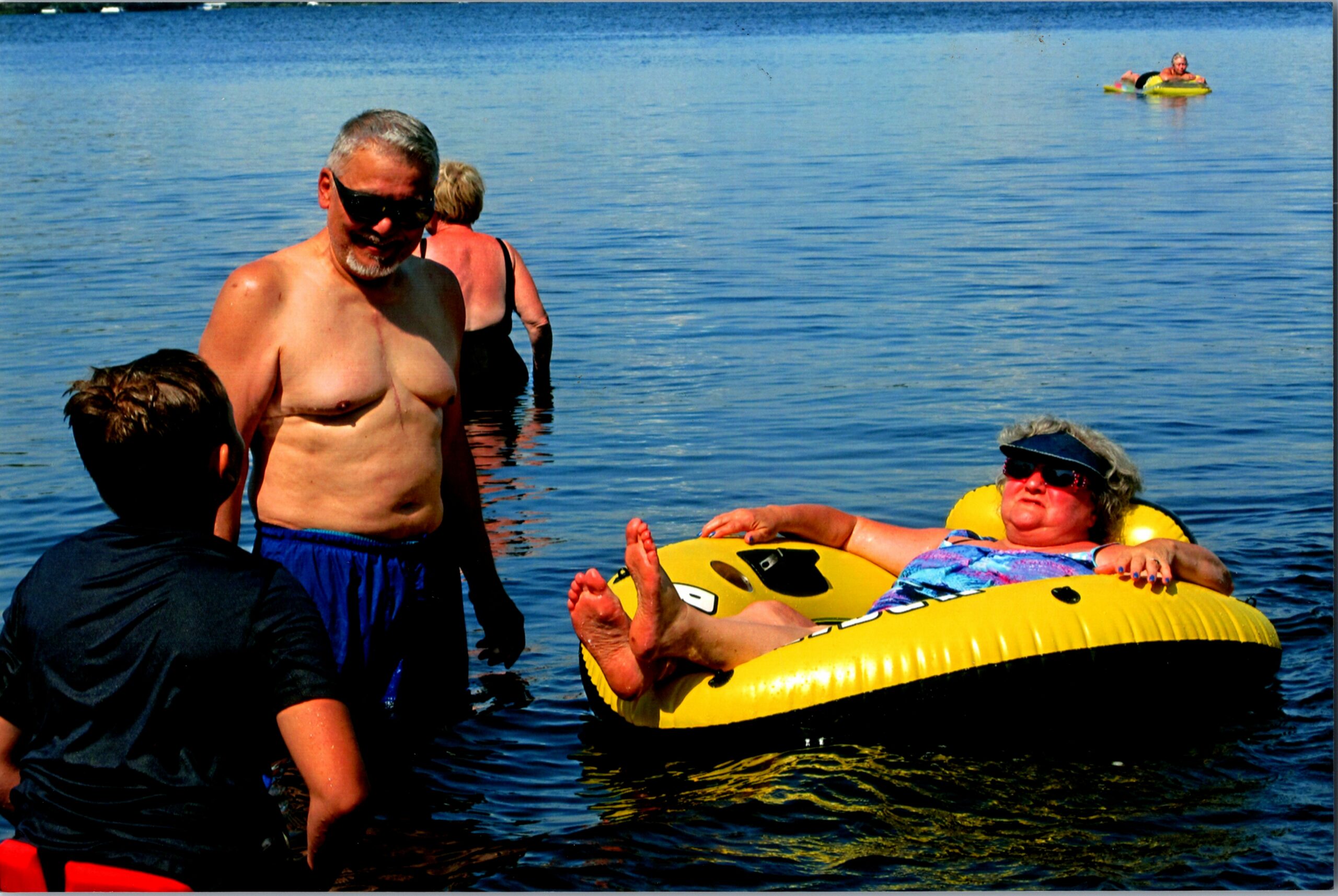 Group of people in a lake; a shirtless man wearing sunglasses stands in shallow water while others swim nearby, a yellow inflatable tube is present too, with a person resting on it on the right.